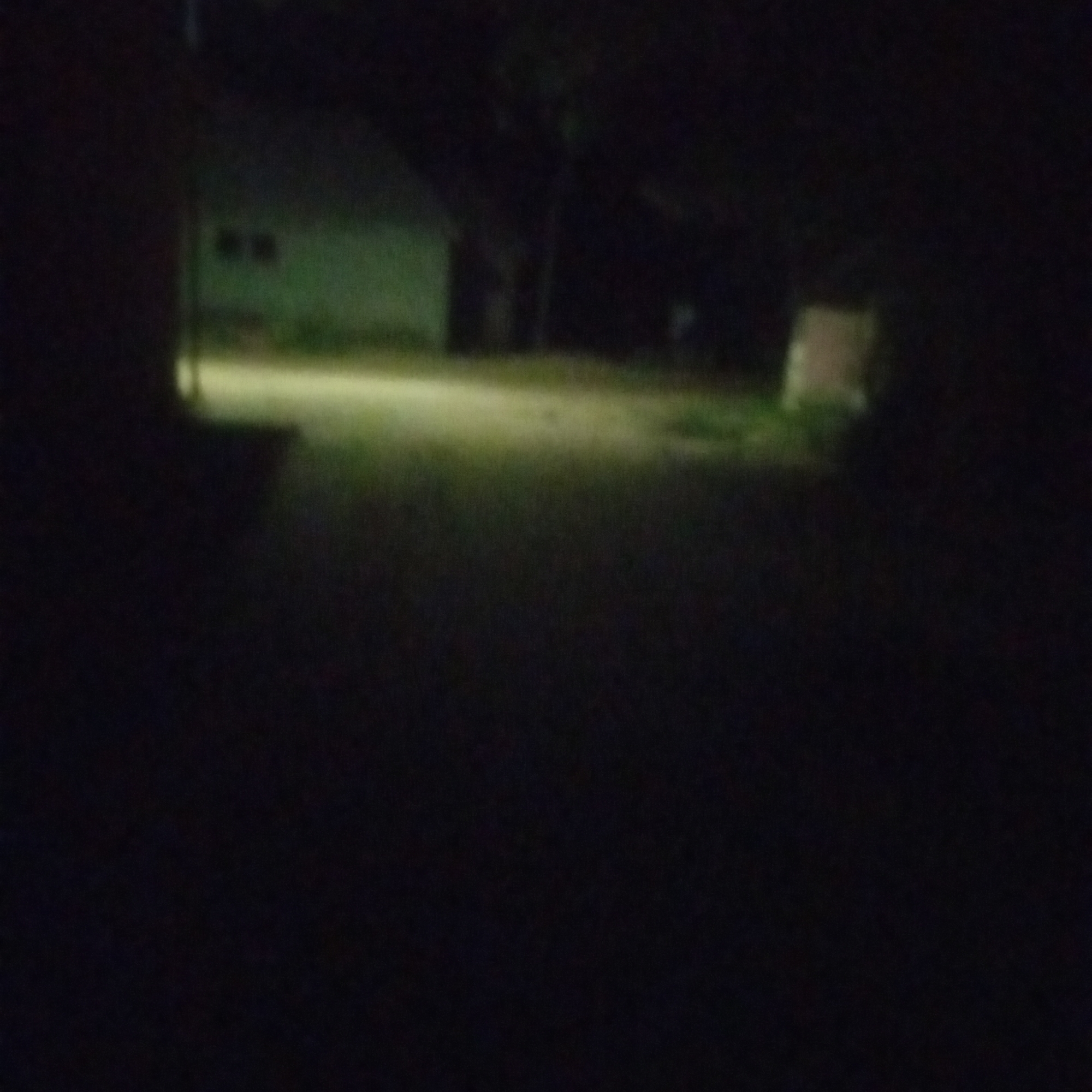 The image depicts a dimly lit outdoor scene, likely taken at night, in Jalingo, Nigeria. A pale, yellowish light illuminates a portion of a dirt or unpaved road in the foreground, with darker shadows on either side. In the mid-ground, a low-lying structure, possibly a building or wall, is visible on the left, painted a light green or off-white. Two dark rectangular shapes, which could be windows, are present on the side of this structure. To the right of the road, a darker, undefined shape suggests the presence of trees or dense foliage. Further to the right and slightly back, a lighter, roughly rectangular object stands out, appearing as a box or a piece of discarded material. The overall impression is one of a rural or semi-rural area experiencing low light conditions.