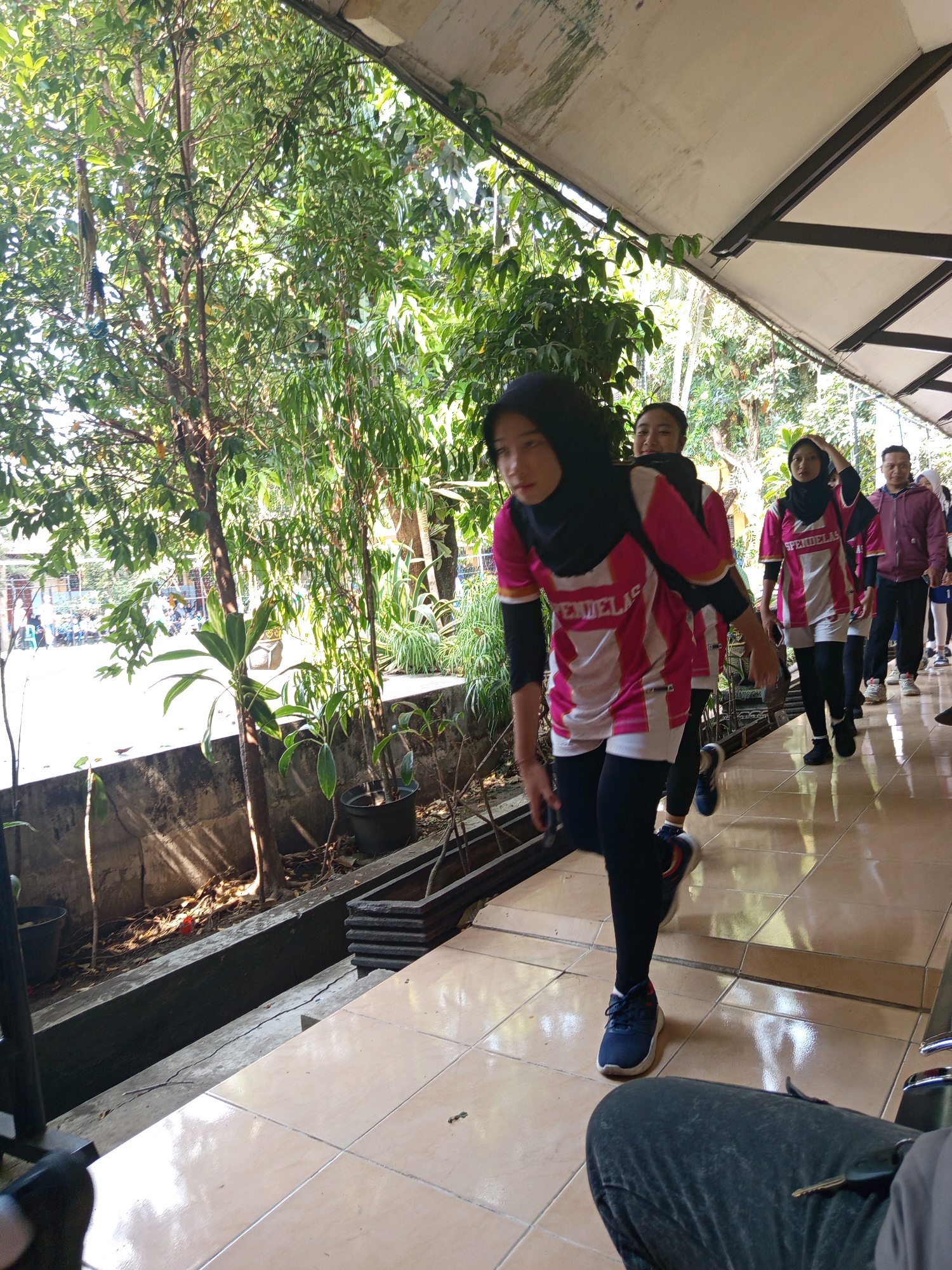 The image depicts a group of young women and a man in a line, walking outdoors in what appears to be a school or community setting in Lesanpuro, Indonesia. The foreground is dominated by the tiled walkway and a person's legs in dark pants, partially obscuring the view. Several young women in matching pink and white sportswear, some wearing hijabs, are in mid-stride. They are all facing the same direction, suggesting they are participating in an activity like a procession or exercise routine. Behind them, a man in a purple jacket and a white shirt is also walking. The environment includes lush green trees and plants bordering the walkway, with a shaded awning overhead. Sunlight filters through the foliage, creating dappled light on the ground. In the background, there are hints of buildings and other people, suggesting a populated area. The overall impression is one of an organized outdoor event.