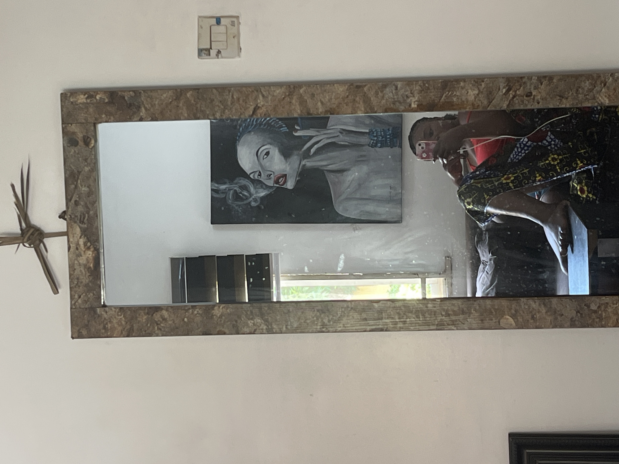 The image depicts an indoor scene, likely a living room, within a residential setting in Abuja, Nigeria. A large, distressed wooden-framed mirror is the central focus. Reflected in the mirror is a woman sitting on a low surface, possibly a bench or stool. She is wearing a red tank top and a patterned skirt. She is holding a smartphone and appears to be taking a selfie. To the left of the woman's reflection, a painting of a woman smoking is visible. The painting is in shades of gray and black with red lips and blue jewelry. Behind the painting, a window with louvred blinds and a view of greenery can be seen. On the wall to the right of the mirror, a white electrical socket with three switches is mounted. The overall ambiance of the room appears casual and personal.