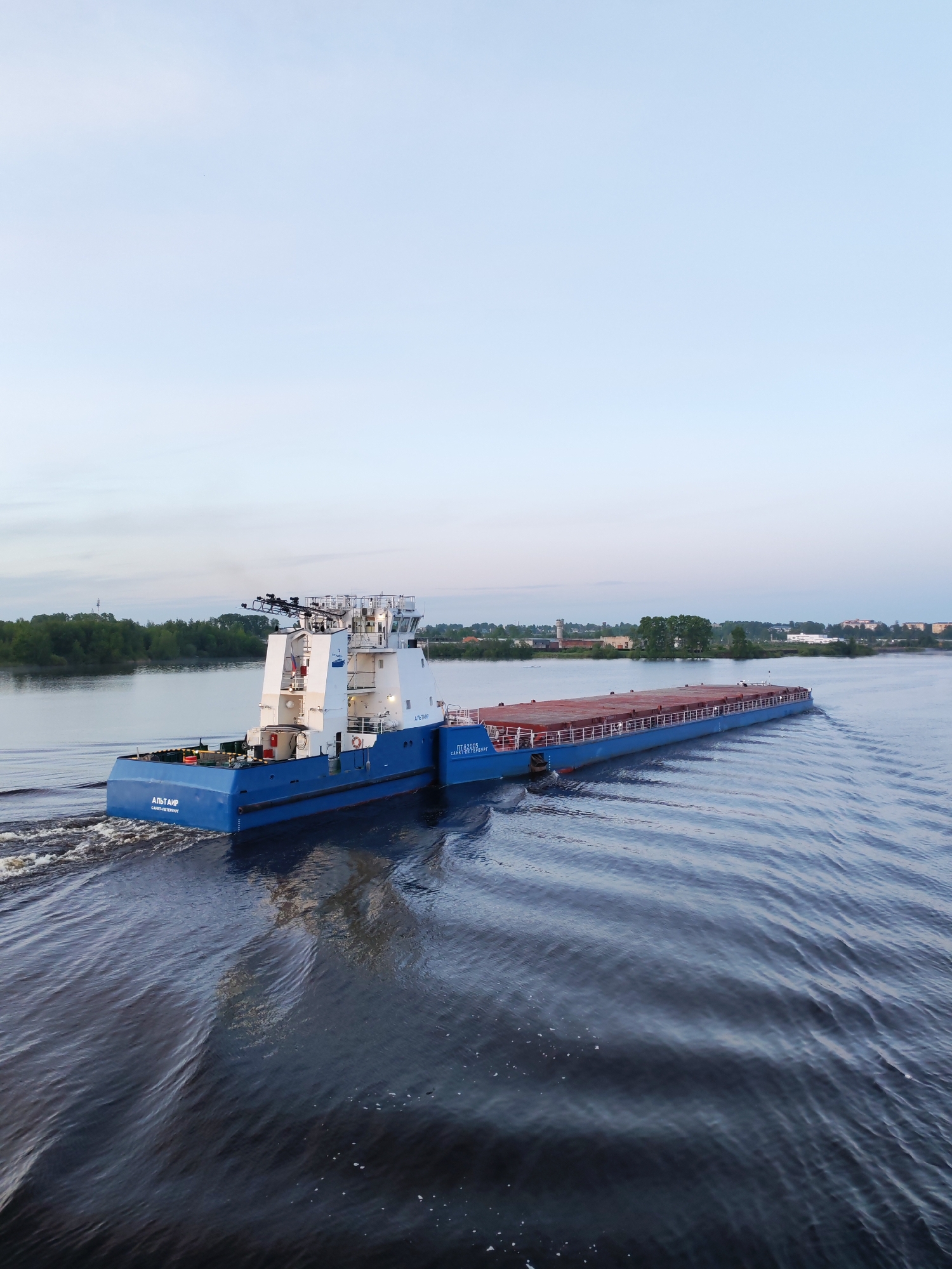 A blue and white barge named "Al'tair" is navigating a river with calm, rippled water. The barge is carrying a long, flat cargo container. The barge is creating a wake as it moves through the water. In the distance, there is a shoreline with trees and buildings. The sky is light blue.