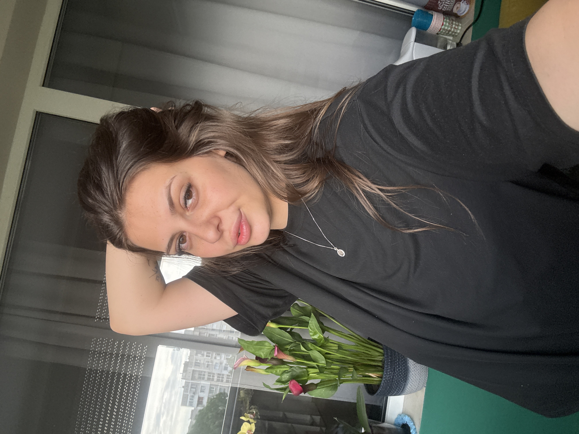 A young woman is posing for a selfie indoors, likely on a balcony or near a window, with a city view in the background. She has long brown hair, which she is holding up with her right hand. She is wearing a black t-shirt and a silver necklace. Her makeup includes subtle eyeshadow and lip gloss. To her left, a potted plant with large green leaves and pink calla lilies is visible. The background shows buildings and what appears to be a cloudy sky. The overall impression is casual and relaxed. The location is not specified in the image.