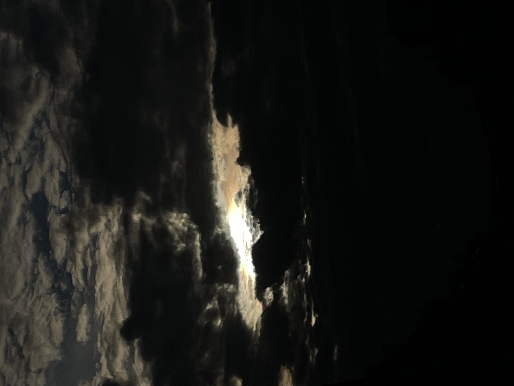 A low-angle, full shot captures a dramatic sky filled with dark, ominous clouds. A sliver of bright light pierces through a gap in the clouds, illuminating the edges with a golden hue, suggesting a sunset or sunrise. The majority of the sky is in shadow, creating a stark contrast between light and dark. The lower portion of the image is entirely black, obscuring any ground-level details or potential city elements, making the location of Unknown, Unknown impossible to ascertain.
