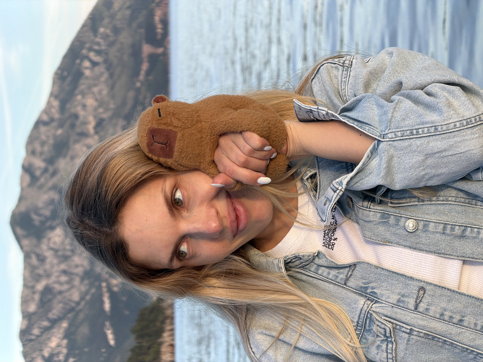 A woman with light brown hair and green eyes holds a brown stuffed bear against her head. She is wearing a white shirt under a light blue denim jacket. The background shows a blurred landscape with mountains and water. The location appears to be near Budva, Montenegro, given the landscape.