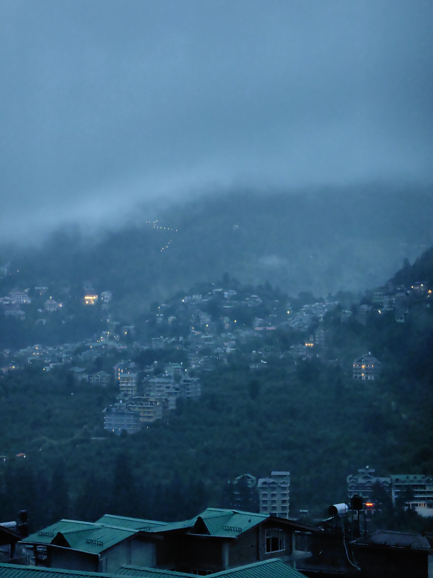 This is a nighttime, aerial view of a densely populated hillside in Vashisht, India. The scene is dominated by a thick, ethereal fog that blankets the upper portion of the image, obscuring the sky and a distant ridge. Below the fog, the slopes are covered with a dense array of buildings, many of which are illuminated with warm, yellow lights, suggesting residential or hotel accommodations. The architecture appears varied, with some structures featuring multiple stories and balconies, while others are smaller and more spread out. Some buildings are topped with flat, green roofs. In the foreground, closer to the viewer, more buildings are visible, also with green roofs, some of which have water tanks on top. The overall impression is of a vibrant, active community nestled within a mountainous landscape, shrouded in the mystical atmosphere of a foggy night.