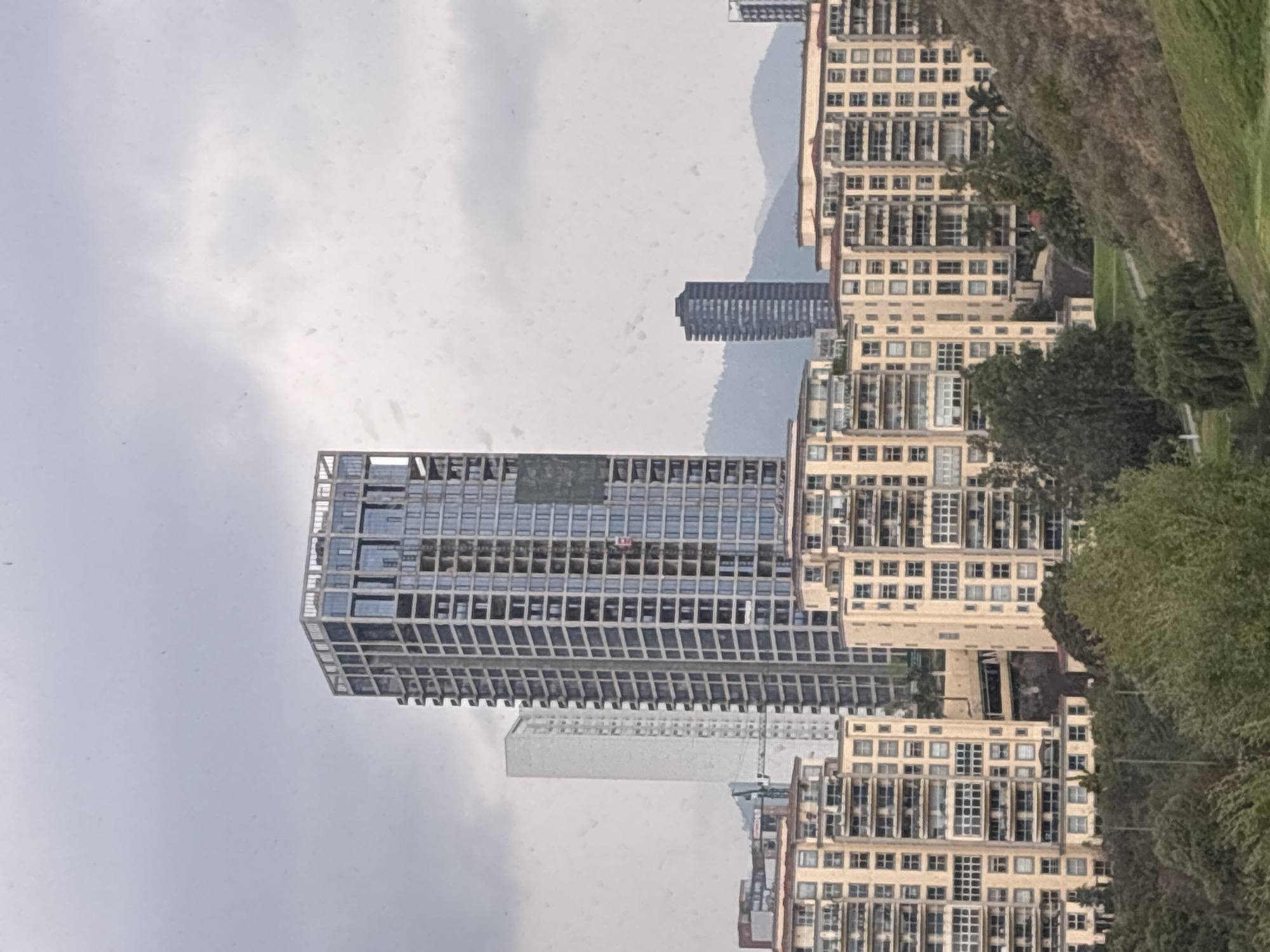 The image shows a cityscape in Huixquilucan de Degollado, Mexico. Prominent in the view is a modern, high-rise building under construction, its skeletal frame visible against a cloudy sky. Adjacent to the construction site are several completed apartment buildings with repetitive window patterns. Vegetation, including trees and grassy areas, flanks the right side of the image. Further in the background, a shorter, darker skyscraper is partially obscured by the sky. The overall scene depicts a mix of urban development and natural elements under overcast weather conditions.