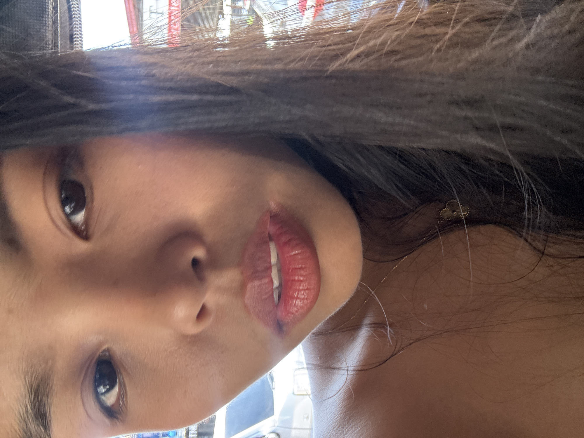 This rotated close-up portrait captures a section of an individual's face, likely a young woman, with her head oriented sideways as if the image was taken with the camera rotated 90 degrees clockwise. Her long, dark brown hair frames the upper part of the frame.
The subject's dark brown eyes are open and directed towards the camera. Her nose is visible in the center, and her lips, adorned with a reddish-pink lipstick, are slightly parted, revealing some teeth. A distinct dark mark or smudge is present near the right corner of her mouth, which could be smudged lip liner or a small blemish.
A small, gold-colored accessory is visible within her hair strands on the right side of the image, possibly an earring or a hair ornament. The background is brightly lit, blurred, and largely overexposed, indicating natural daylight conditions, likely outdoors or near a window. The lack of distinct features in the background prevents identification of specific location cues, though the context provided places this scene in Davao City, Philippines. No visible text is present in the image. The overall impression is a personal close-up selfie taken during the daytime.