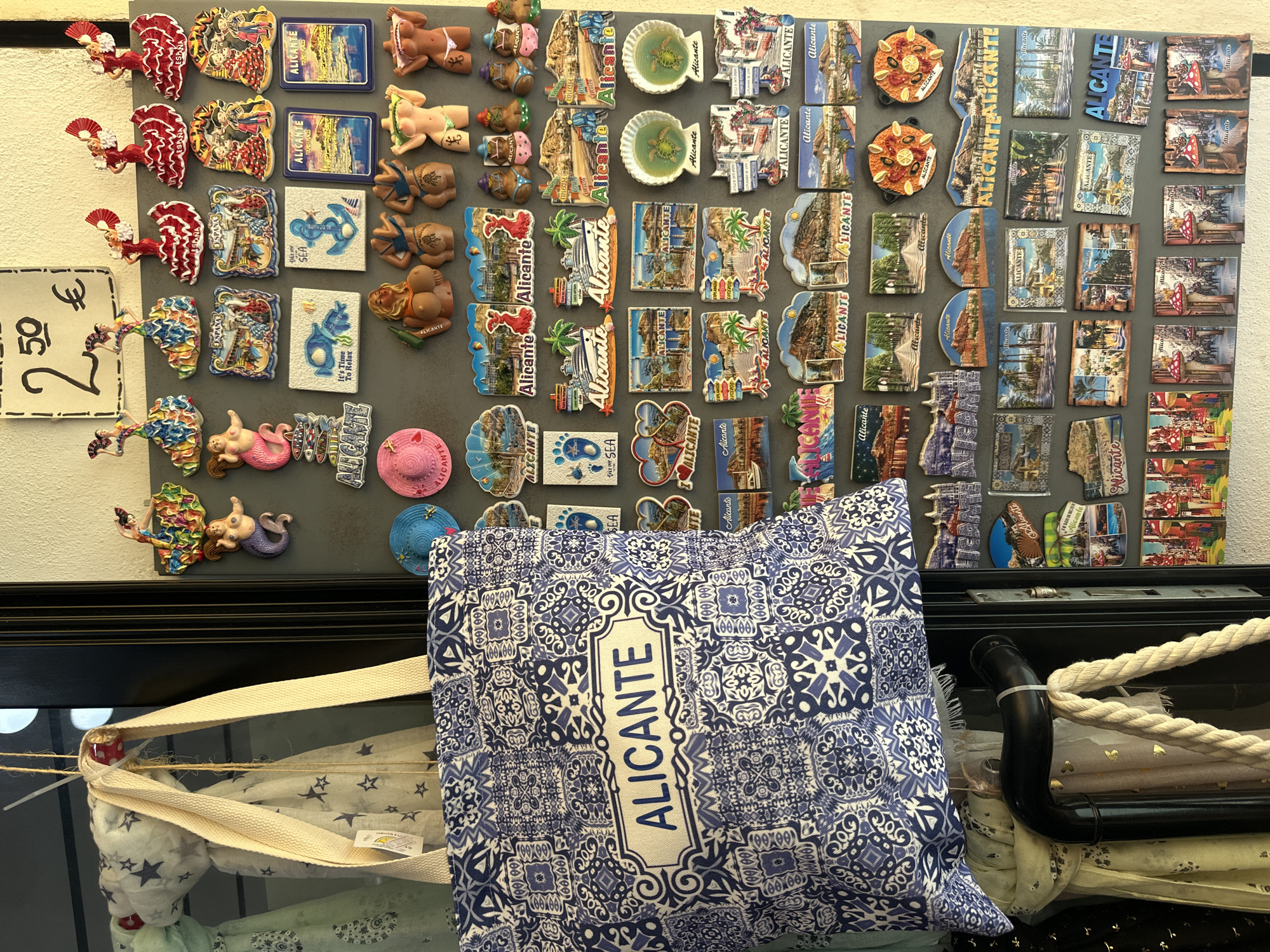 The image displays a vibrant collection of souvenir refrigerator magnets attached to a gray metallic surface, likely a display board in a shop or market. The magnets vary widely in shape, size, and design, often featuring bright colors.
Prominent subjects include:
* Flamenco dancers in red dresses and fans.
* Figurines of people in swimwear, some with exaggerated features.
* Mermaids with colorful tails.
* Miniature food items, specifically paella dishes with seafood and lemon wedges.
* Small decorative hats, some pink, some blue.
* Scenic depictions of cityscapes, landmarks, beaches, and palm trees, typical of a coastal tourist destination.
* Abstract patterns and designs, including some with turtles in shell-shaped dishes.
Repeated text on many magnets, and a blue and white patterned tote bag in the foreground, reads "ALICANTE" or "Alicante," strongly indicating the location is Alicante, Spain. Other visible text includes "ESPAÑA" (Spain), "It's Time To Relax," and "take me SEA."
A white price tag on the left side shows "2.50 €", suggesting the price of items, likely the magnets. The tote bag, featuring a blue and white tile-like pattern with "ALICANTE" in a central scroll, partially obscures the lower part of the magnet display. Below the display, through a transparent surface, glimpses of folded textiles, possibly scarves or umbrellas, are visible, secured with ropes and white zip ties. The overall scene suggests a retail setting, likely a tourist shop, offering local souvenirs.