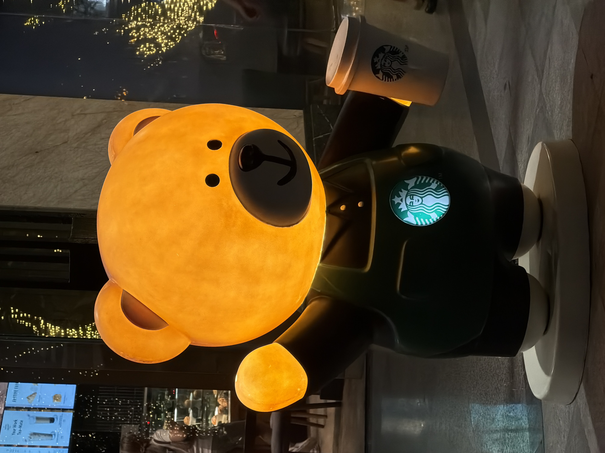 This is an eye-level shot of a large, illuminated teddy bear statue dressed in a Starbucks uniform. The statue is orange and glows from within, with black eyes, nose, and mouth. It wears a dark green apron with the Starbucks logo prominently displayed on the chest. The bear is holding a Starbucks coffee cup in its right hand and its left arm is raised with a glowing orange orb at its fingertip. The statue stands on a white circular base. In the background, out-of-focus, there appears to be a building with illuminated signage and decorative lights, possibly a storefront. There are also reflections on a glass surface in the background, and the floor is tiled. The setting is likely a public area or an exterior of a commercial establishment, such as a Starbucks branch. The image does not provide any specific visual cues to place it in Nha Trang, Vietnam.