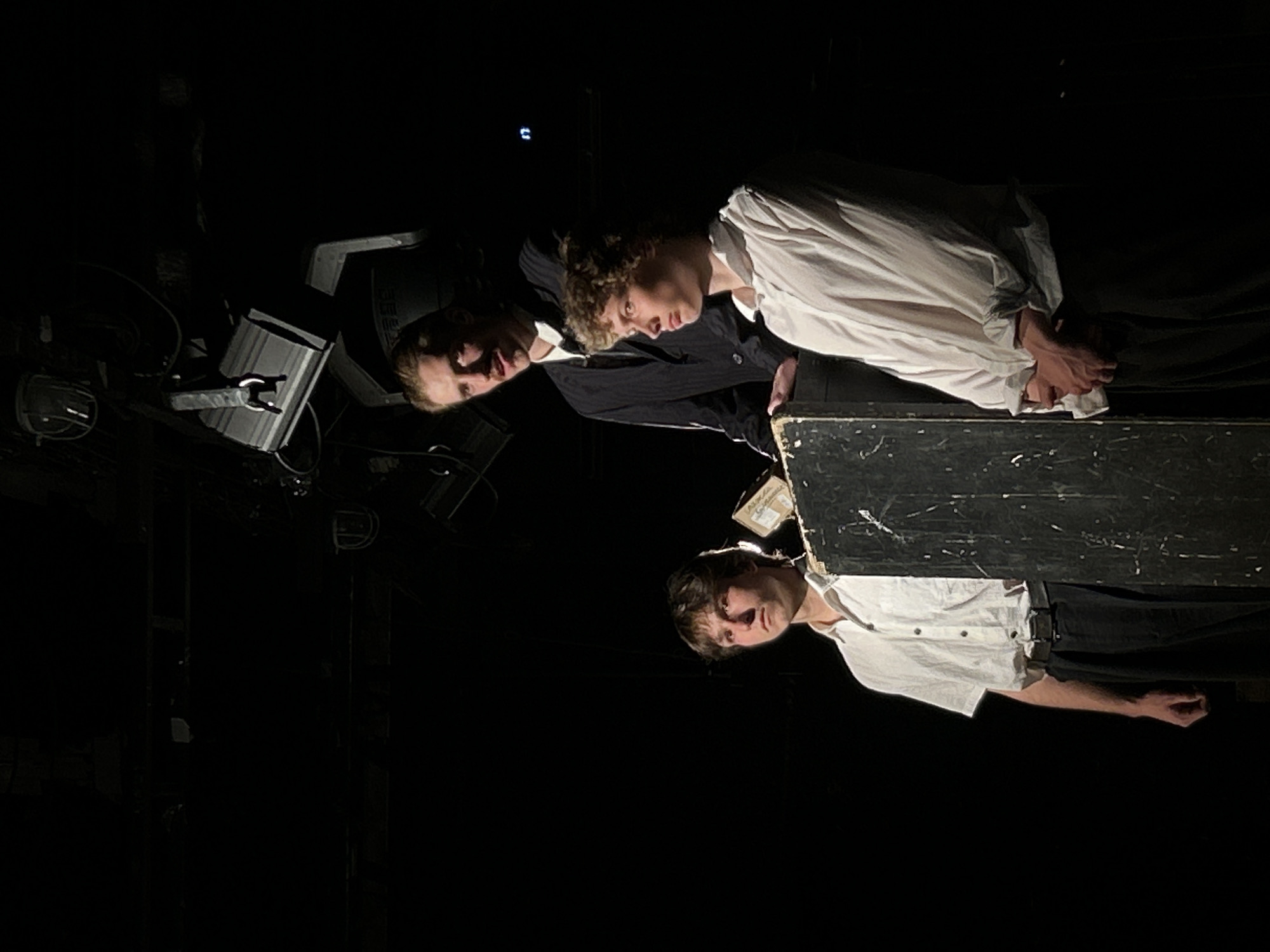 The image depicts three individuals on a stage in a dimly lit environment in Unknown, Unknown. Two of them are wearing white shirts, while the third one is in a black jacket. They are positioned around a dark, wooden object that appears to be part of the stage setup. To the left, there are some equipment pieces, possibly lighting or sound equipment. The overall scene suggests a theatrical or performance setting.