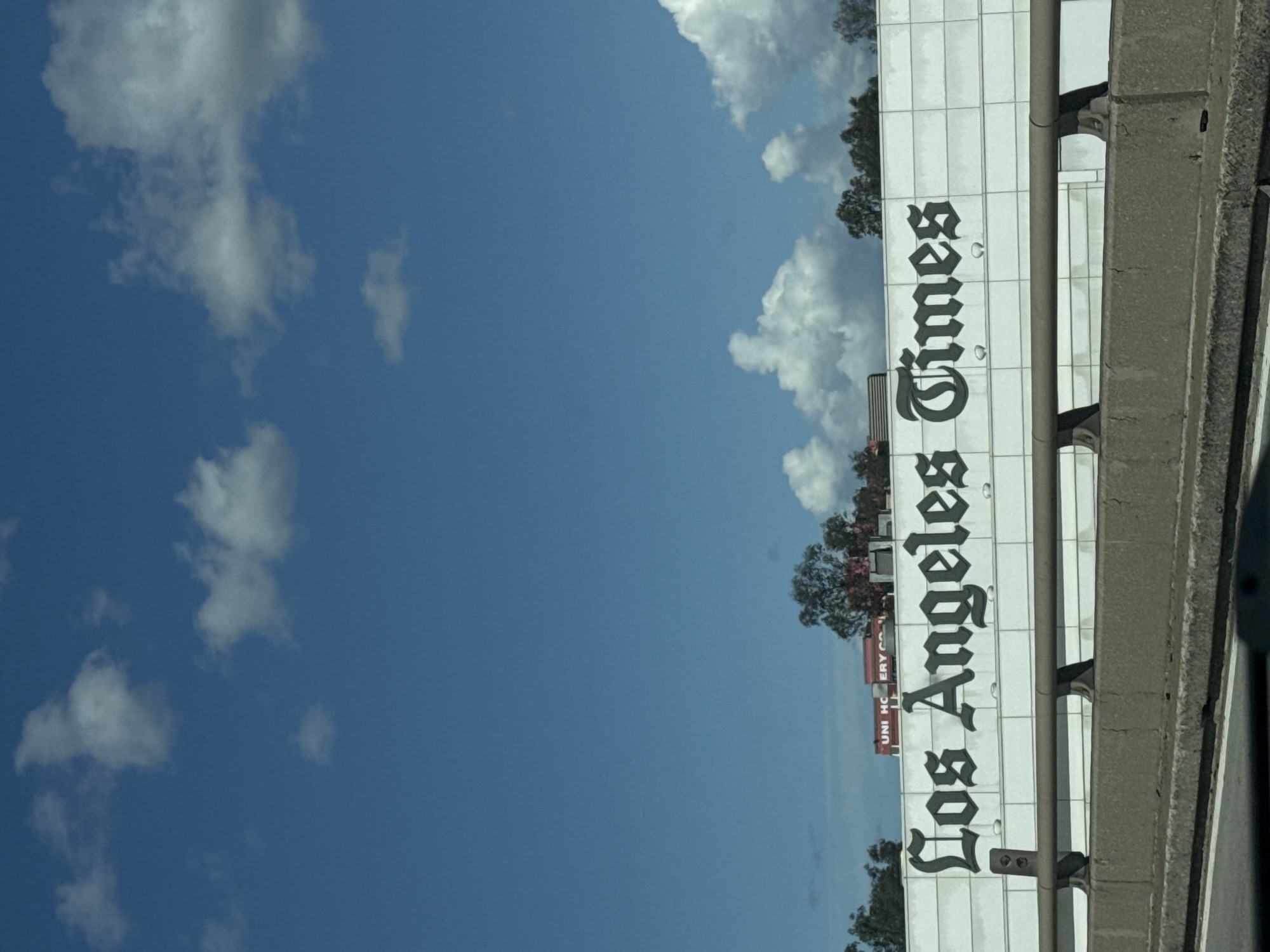 The image depicts the Los Angeles Times building in Los Angeles, United States of America. The building's sign is visible, showcasing the name "Los Angeles Times." The sky is blue with some clouds. A highway runs next to the building. A small Uni-Kool banner is visible behind the sign. The image appears to have been taken from a moving vehicle.