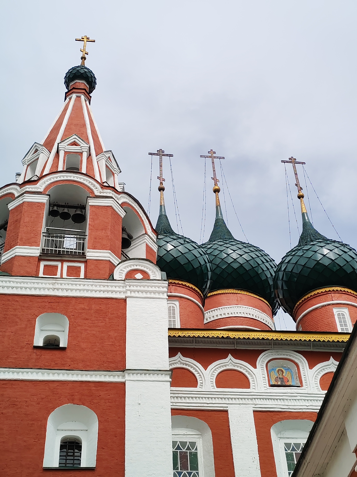 The image displays the exterior of a red brick church in Rusanovo, Russia. The architecture features prominent white decorative trim and multiple green, onion-shaped domes topped with crosses. A bell tower with bells is visible on the left side of the image. The sky above is overcast. The scene is a religious building, likely a Russian Orthodox church, given the architectural style and the presence of crosses on the domes. The image is a medium shot, focusing on the upper portion of the church structure.