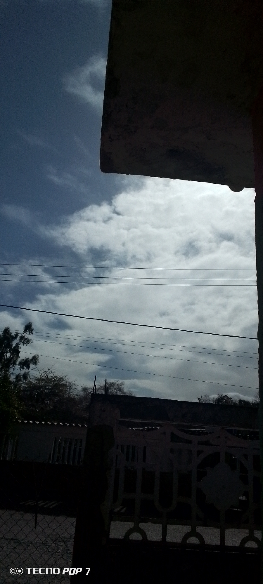 The image shows a view of a sky with scattered clouds, electrical wires, and partial views of structures and foliage. A corner of a building is visible in the upper right, casting a shadow. Below, a section of a white fence is partially visible, along with a glimpse of a building behind it. Some trees are also seen. The picture was taken in the city of Altagracia, Venezuela.