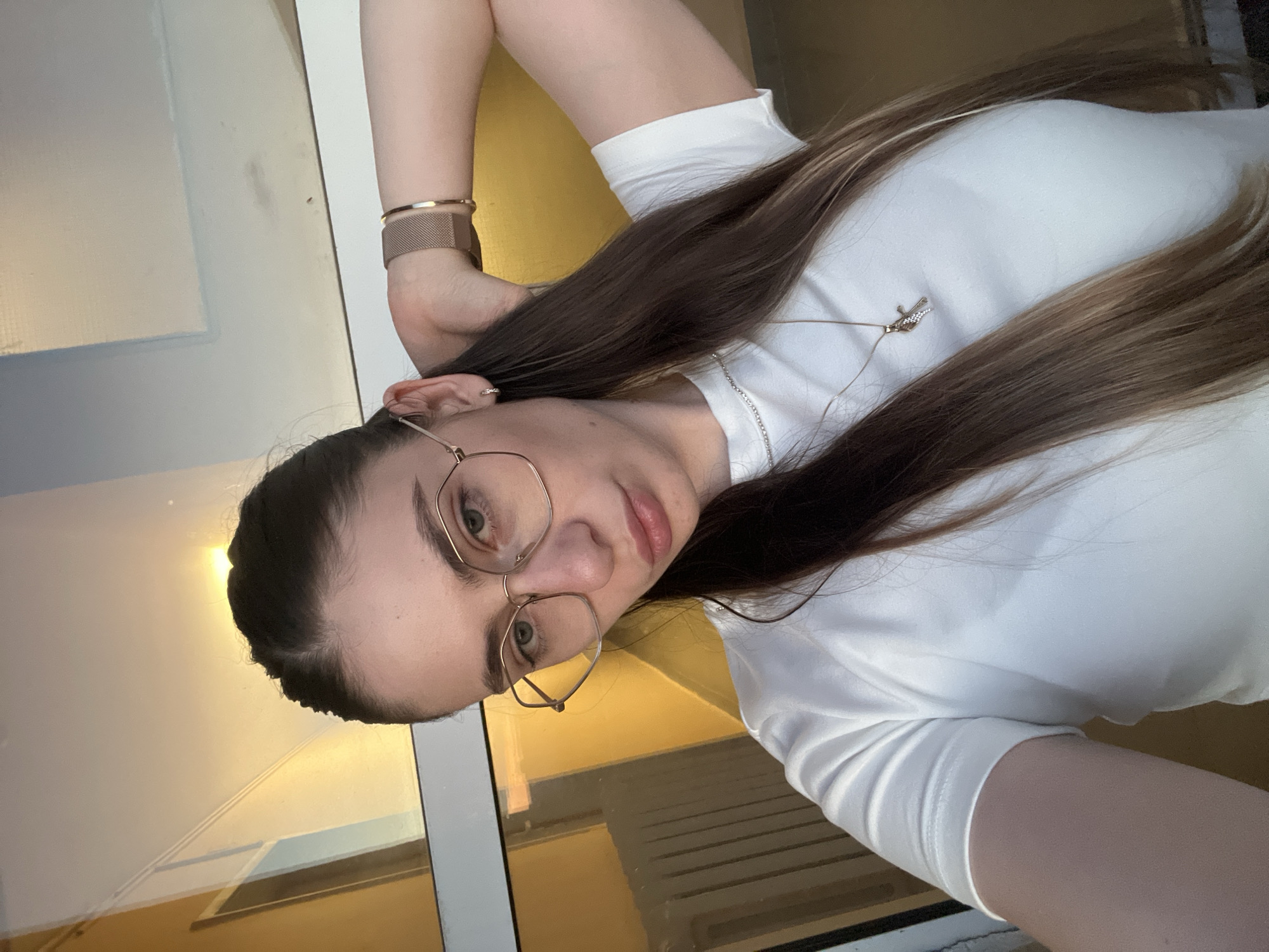 A young woman with long, dark brown hair, wearing glasses and a white t-shirt, poses for a selfie. She is leaning against a large window with a yellow tint. Her right arm is raised behind her head, and she is looking down and to the left. She is wearing a rose gold watch on her left wrist and a delicate necklace with a cross pendant. The background appears to be an indoor space with a radiator visible through the window. The lighting suggests it is either late afternoon or early morning. The location is unknown.