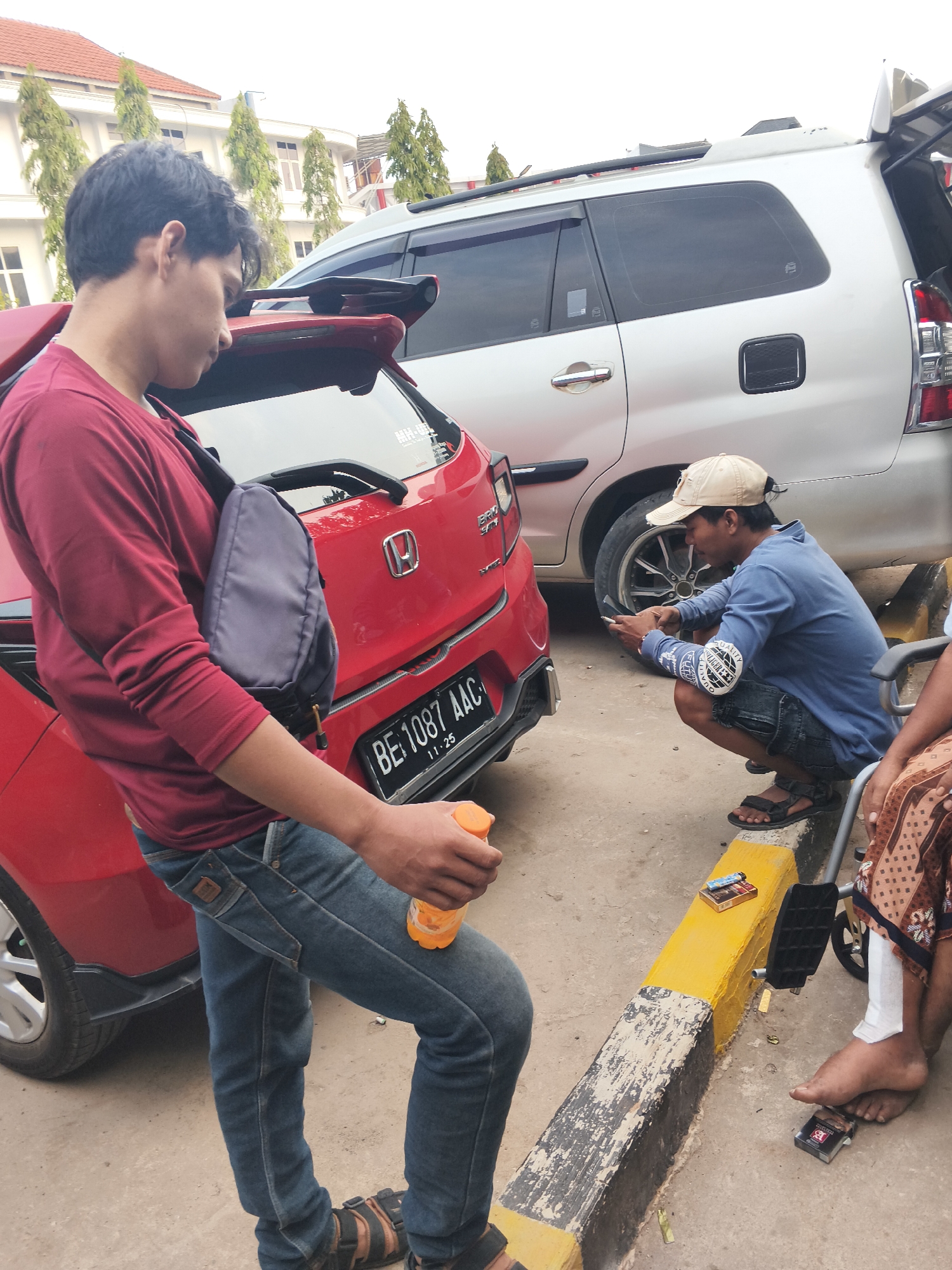 In Bandar Lampung, Indonesia, two men and a partially visible woman are gathered near two cars. A red Honda Brio is parked to the left, facing right, with its rear partially visible. To its right is a silver Toyota Innova, facing left, with its rear tailgate open.
The first man, wearing a maroon long-sleeved shirt and blue jeans, stands to the left, holding an orange bottle in his right hand. A grey sling bag is worn across his chest. He is looking downwards.
The second man, wearing a blue long-sleeved shirt, dark shorts, a beige cap, and sandals, squats on the right side of the image. He is looking at his phone, which he holds in his left hand. There are patches on his sleeves.
A woman, her legs and feet partially visible, sits in a wheelchair on the far right. She wears a patterned skirt. Her right foot is bare, and there is a bandage on her lower right leg. A pack of cigarettes is on the ground near her feet.
A yellow-and-black painted curb runs across the foreground. Two lighters and a pack of cigarettes are placed on the yellow section of the curb. The ground is paved with concrete. In the background, a white building with red-tiled roofs and several trees are visible.