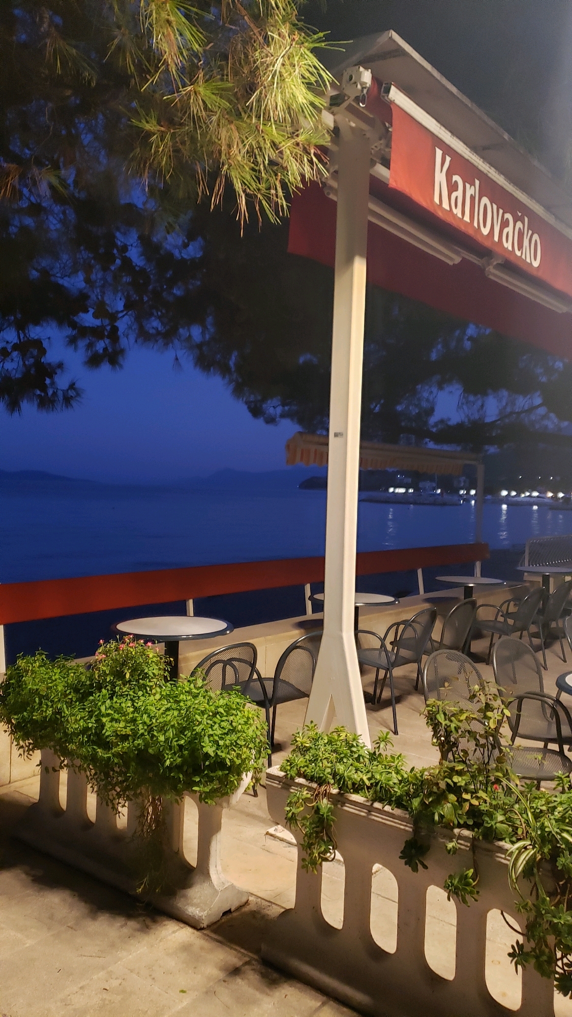The image shows an outdoor seating area of a cafe or restaurant, possibly in Općina Tučepi, Croatia, during twilight. The scene is illuminated by artificial lighting, creating a warm glow on the outdoor furniture and the surrounding vegetation. In the foreground, there are several white concrete planters filled with lush green plants, some of which hang over the sides. Behind the planters, rows of dark metal chairs and round tables are arranged, suggesting a dining or seating area. A large white support pole is prominently positioned in the center of the frame, holding up a red awning with the text "Karlovačko" printed in white letters. In the background, a calm body of water, likely the Adriatic Sea, stretches out towards the horizon. Mountains are visible in the distance, softened by the evening haze. The sky is a deep blue, indicative of the transition from day to night. Pine tree branches hang into the frame from the upper left corner, their needles illuminated by the artificial lights. The overall atmosphere is serene and inviting, typical of a seaside establishment in the evening.