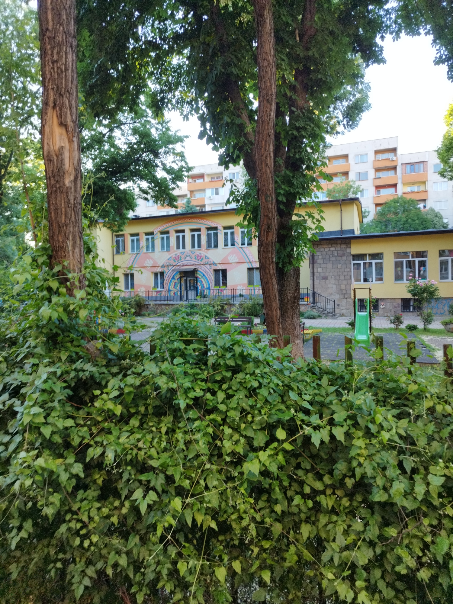 The image shows a yellow, two-story building, possibly a kindergarten or school, situated in a green, park-like setting. A multi-story residential building is visible in the background. In the foreground, a dense hedge or vegetation obscures the view, with trees framing the building. A green slide and playground equipment are partially visible to the right. The building features decorative archways, white windows, and a stone exterior section. The location is likely in Sofia, Bulgaria, judging from the architectural style of the buildings. There are no people visible in the frame.