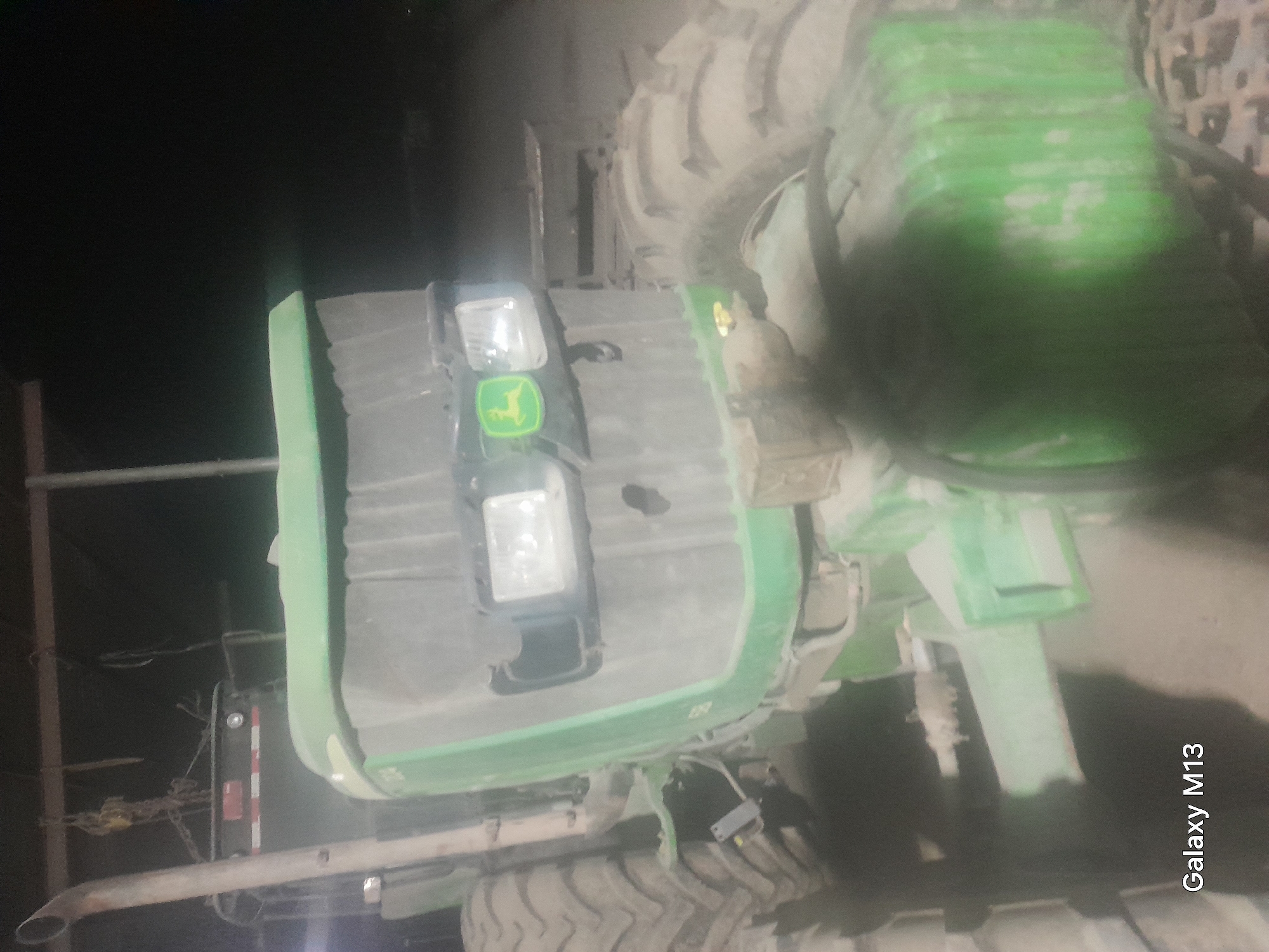 A close-up, nighttime photograph shows the front and right side of a green John Deere tractor. The tractor appears dusty. The front of the tractor features two rectangular headlights and a prominent John Deere logo. A large, knobby tire is visible on the right side. In the background, dimly lit objects, possibly part of a structure or other farm equipment, are present. The bottom left corner of the image displays the text "Galaxy M13".
