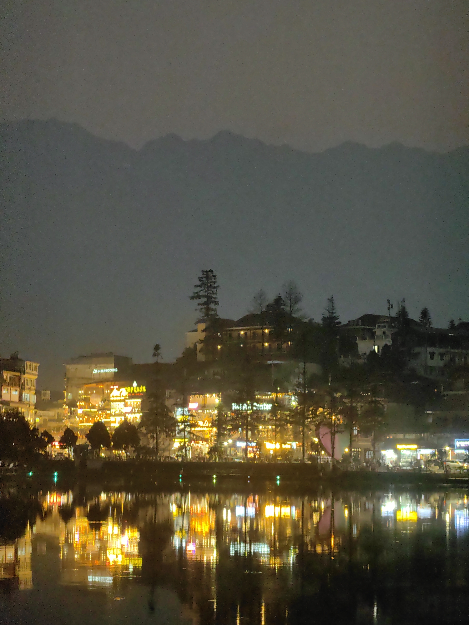 This image depicts a nighttime panoramic view of Sa Pa, Vietnam, specifically overlooking Sa Pa Lake and the town built into the surrounding hillsides.
In the foreground, the dark, still surface of the lake prominently reflects the vibrant artificial lights from the buildings on the opposite shore, creating elongated streaks and pools of yellow, orange, and white illumination. Small green lights are also visible on the water's surface.
The midground features the lake's shoreline, densely populated with multi-story buildings extending up the sloping terrain. These structures are heavily illuminated with bright, predominantly warm-toned lights, indicating commercial activity such as shops, restaurants, and hotels. Identifiable signage includes "INTERBOOKING" on a building towards the left and "Quán" (Vietnamese for eatery) on another prominent structure. Numerous trees, both deciduous and coniferous, are interspersed among the buildings, some appearing as dark silhouettes against the bright facades. While indistinct, human figures can be perceived as dark shapes on the illuminated promenade along the shore, suggesting pedestrian activity.
In the background, a distant mountain range rises, its contours softly defined and largely obscured by low visibility or atmospheric haze, appearing as a dark silhouette against a slightly lighter, overcast night sky. The overall scene is characterized by the diffuse glow of urban lighting under a dark, possibly misty, sky.