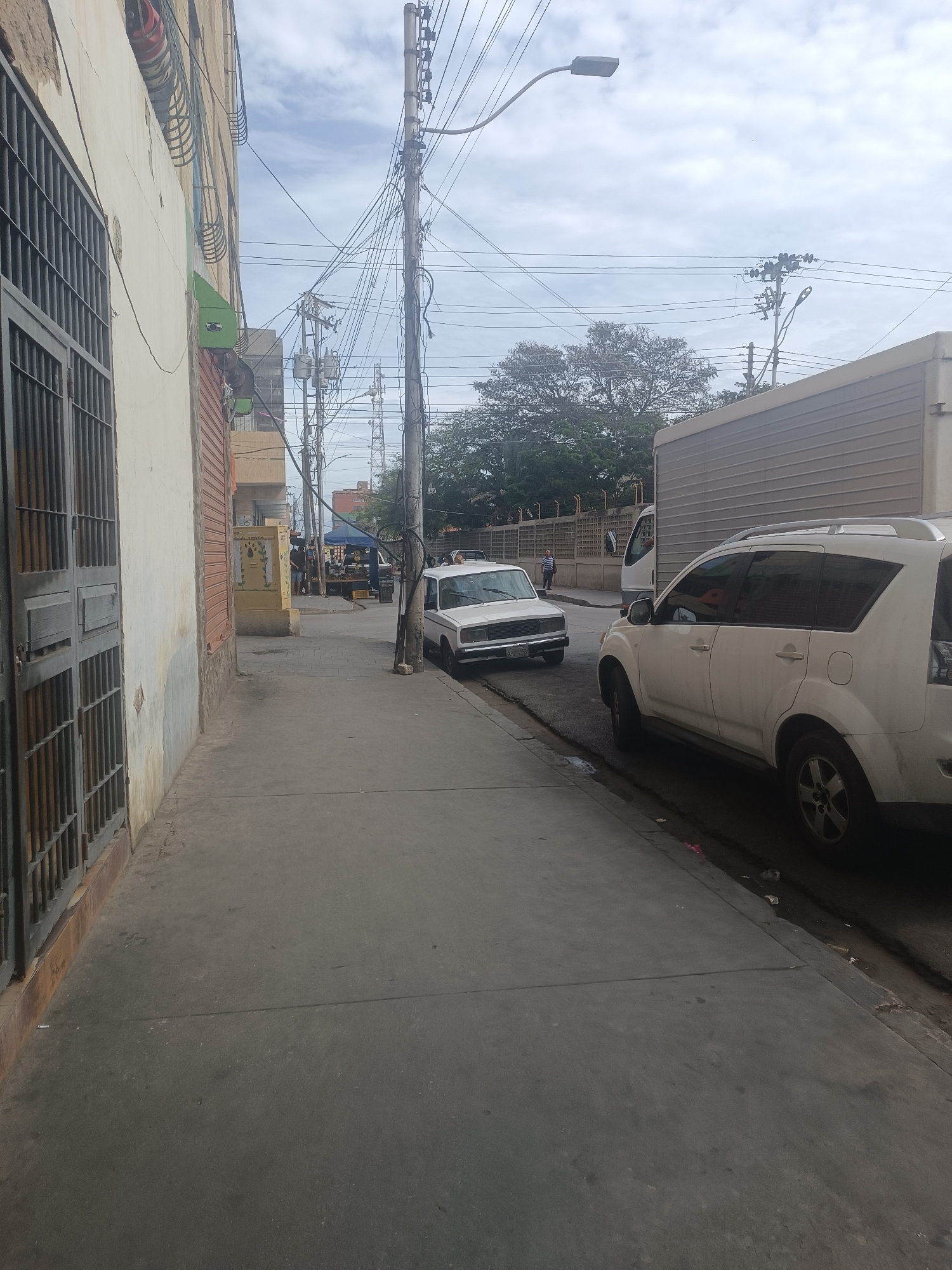 This image depicts an urban street scene in Porlamar, Venezuela, under an overcast sky. The view is from a sidewalk looking down a street.
On the left, a light-colored building with a large, dark-grey metal security gate occupies the foreground. The building's facade shows wear, and exposed utility conduits, some painted green, run vertically along its side. The concrete sidewalk is visible, extending into the distance, with a yellow utility box featuring black markings positioned further down. Beyond this, street vending activity is discernible under a blue canopy.
The street itself runs parallel to the sidewalk, accommodating multiple parked vehicles. A white sedan is parked close to the curb, followed by a white SUV with roof rails, facing towards the camera. Behind the SUV, a large light-colored box truck is parked. The front portion of another white truck is visible between the SUV and the box truck. The curb and gutter line the street, showing some debris and potential standing water.
In the mid-ground and background, numerous utility poles, both wooden and metal, rise from the sidewalk and street, densely interconnected with a complex network of overhead electrical and communication wires. A modern streetlamp fixture extends from one of these poles over the street. Green trees and foliage are visible further in the background, behind a fence. Several individuals are present: one person stands near the white sedan, another walks on the sidewalk near the street vendors, and a third person stands near the fence in the mid-distance on the right.