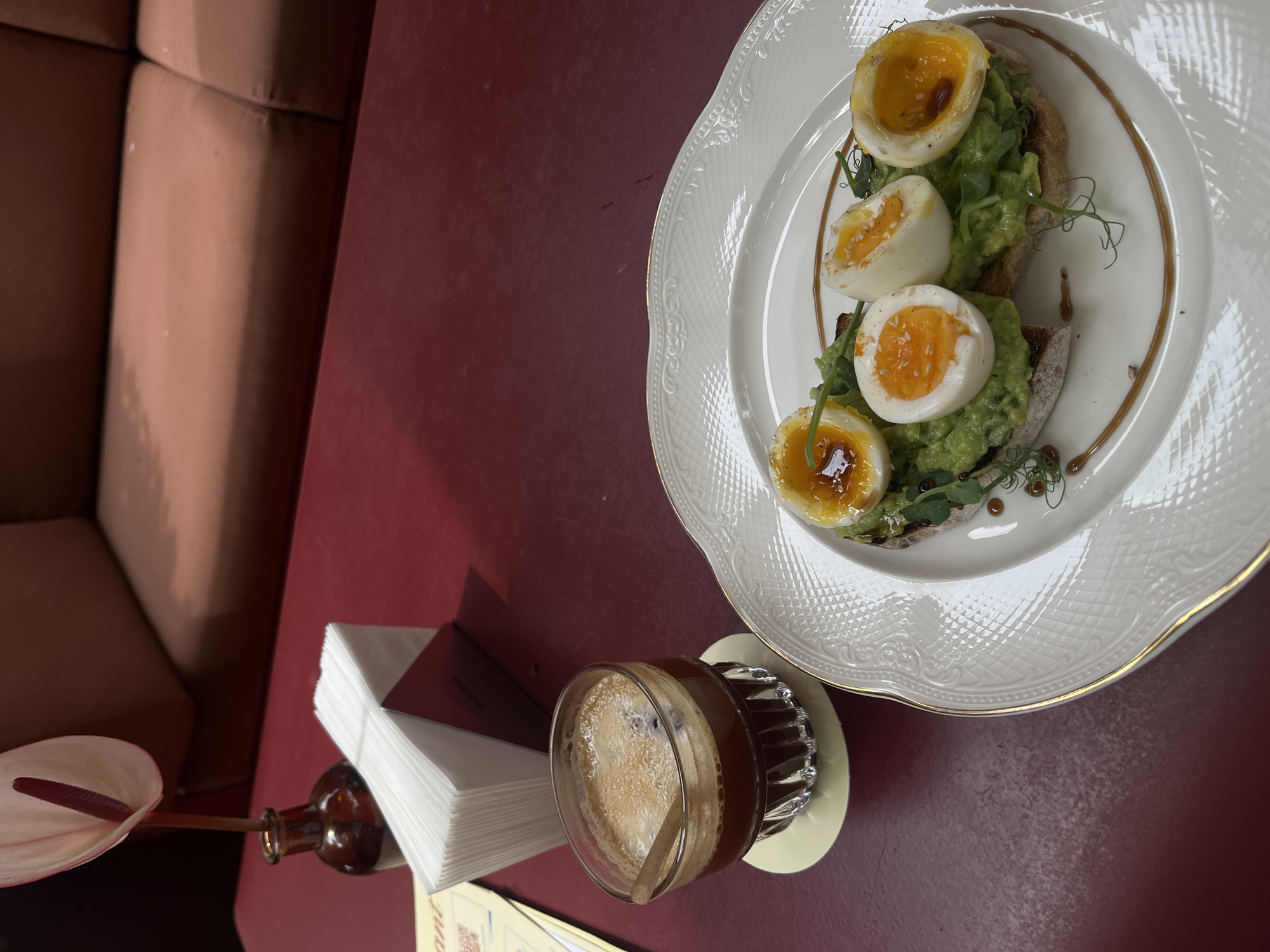 A top-down, slightly tilted shot shows a red table in a restaurant setting. A breakfast plate holds avocado toast topped with three sliced soft-boiled eggs, garnished with sprouts, and drizzled with a dark sauce. Next to the plate, there is a coffee cup on a beige coaster, a stack of napkins, a small amber glass bottle with a single flower, and a corner of a menu. A portion of a tan-colored sofa is visible on the left side of the image. Based on the location, this is likely in Yerevan, Armenia.