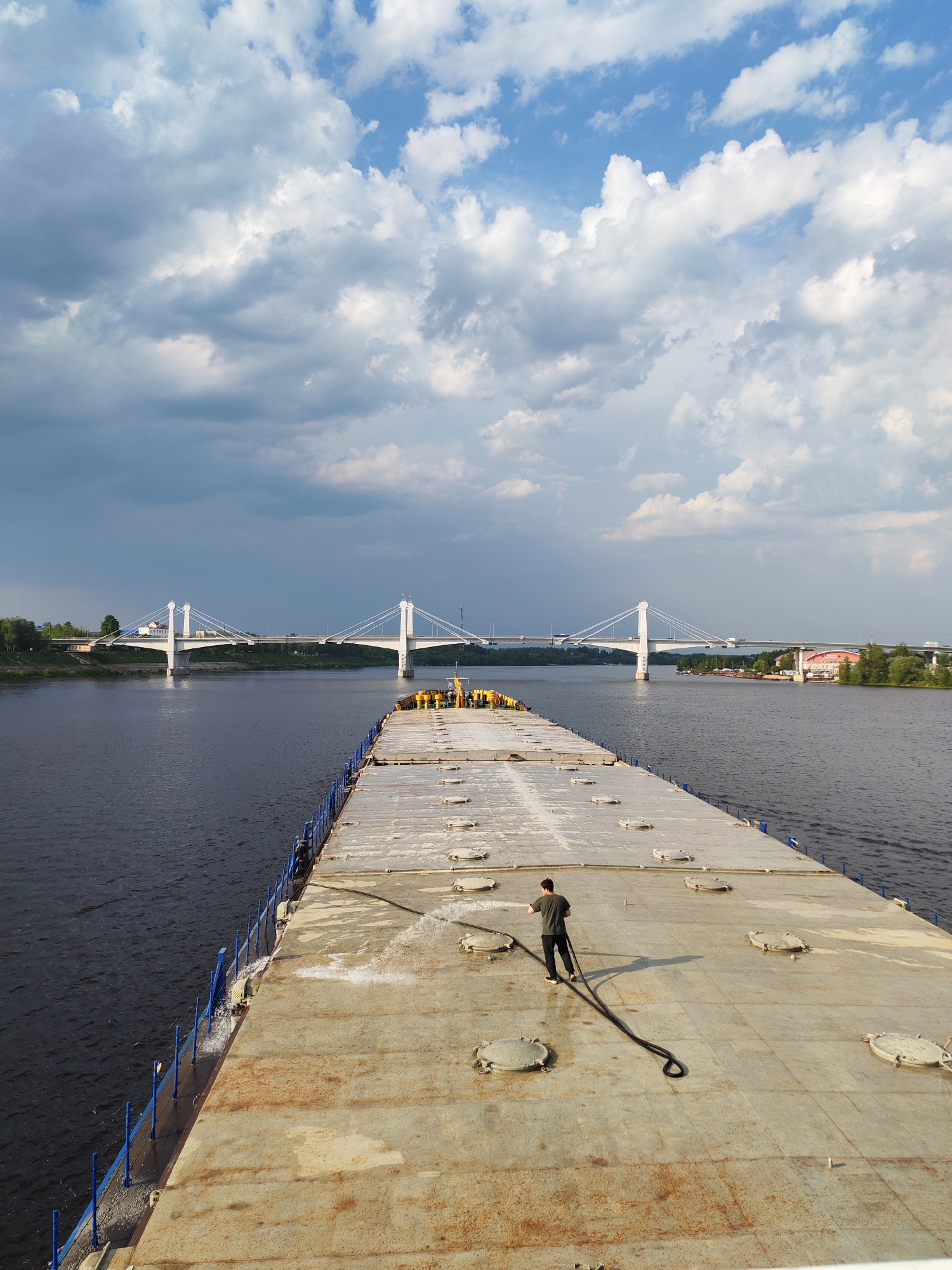 The image captures a wide-angle perspective of a man washing the deck of a barge sailing on the Volga River, with the city of Kimry, Russia, in the background. The barge deck is gray, rust-stained, and marked with round hatch covers. A man, wearing a dark shirt and pants, is seen holding a black hose, spraying water onto the deck. Blue railings line the edge of the barge. In the distance, a white cable-stayed bridge spans the river, with urban buildings and trees visible on the far shore. The sky above is filled with a mix of white and gray clouds, suggesting a partly cloudy day.