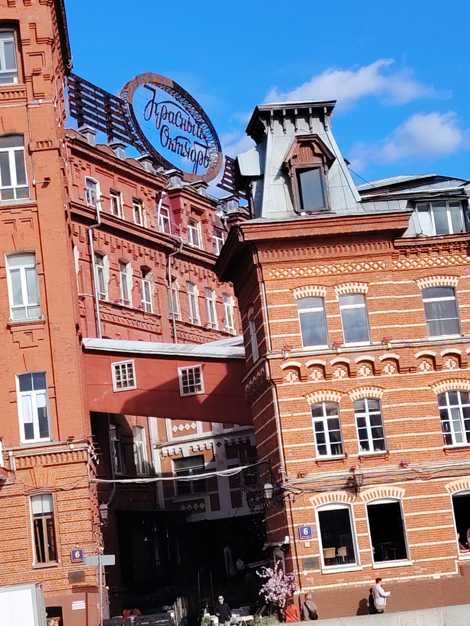 The image shows a close-up view of the red brick buildings of the "Krasny Oktyabr" (Red October) confectionery factory in Moscow, Russia. The ornate architecture features multiple windows with white frames, decorative brickwork, and a distinctive mansard roof. A large oval sign with Cyrillic lettering, "Красный Октябрь", is prominently displayed, suggesting the factory's name. The sky is a clear blue with a few white clouds. Several people are visible in the foreground, some sitting on a low wall and others standing near a light fixture. A small flowering tree adds a touch of color to the scene. The overall impression is of a well-preserved historical industrial complex bathed in daylight.