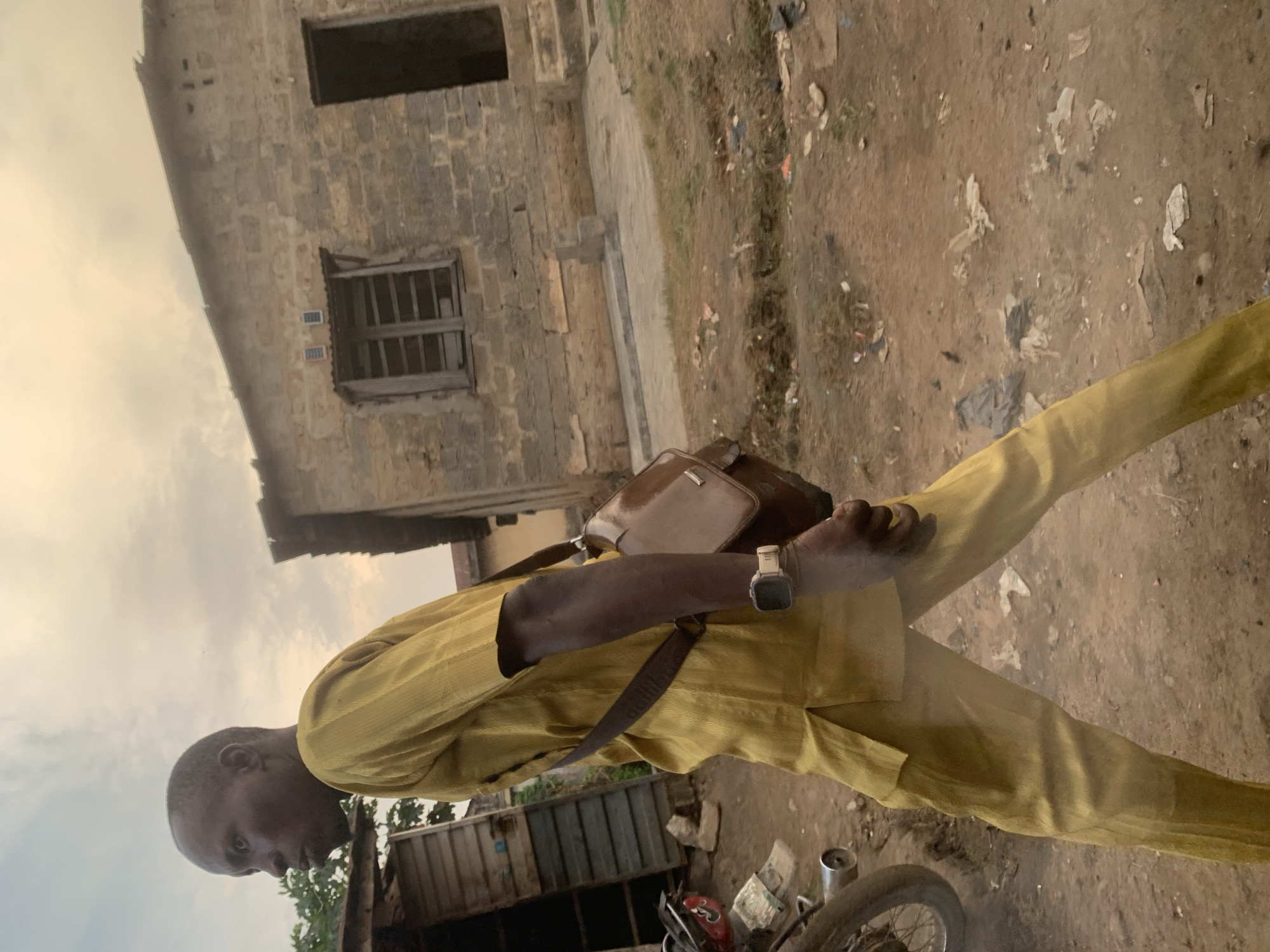 A man in a yellow outfit walks past a dilapidated building and some shacks under a cloudy sky. The man is wearing a yellow top and trousers, a brown messenger bag across his chest, and a watch on his left wrist. He is walking away from the camera towards the right side of the image. To his left, there is a motorcycle with a red seat. In the background, there is a stone building with a broken window and a doorway, and to its left, some wooden shacks. The ground is dirt and littered with debris. The scene appears to be in an urban or semi-urban area in Shagamu, Nigeria.