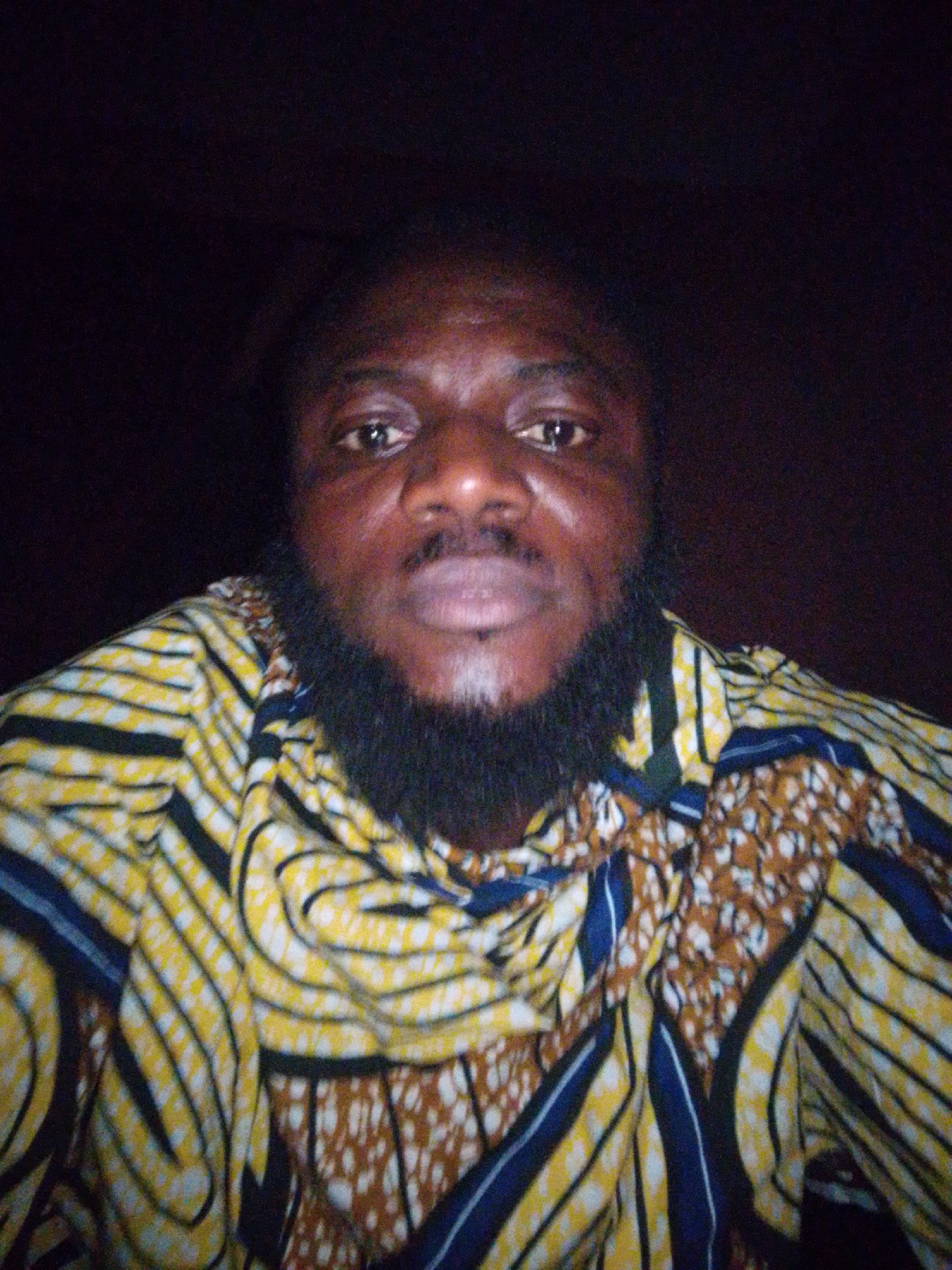 A medium shot shows a dark-skinned man with a full black beard, wearing a colorful patterned shirt. The shirt has yellow and white stripes with blue and brown patterned sections. The background is dark and indistinct, suggesting an indoor setting. The location cannot be confirmed as Lokoja, Nigeria based on the visual information provided.