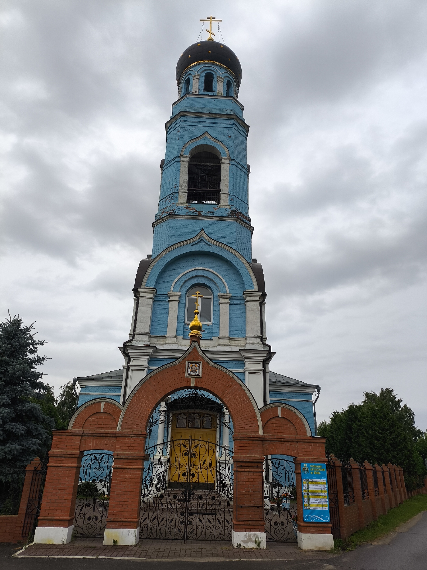 The image shows a blue Orthodox church bell tower with a black dome and a golden cross. The bell tower is made of brick and has arched windows. In front of the church is a red brick gate with ornate black metal fencing. A yellow door is visible through the gate. The sky is cloudy and gray. The church is located in the city of Pokrov, Russia.