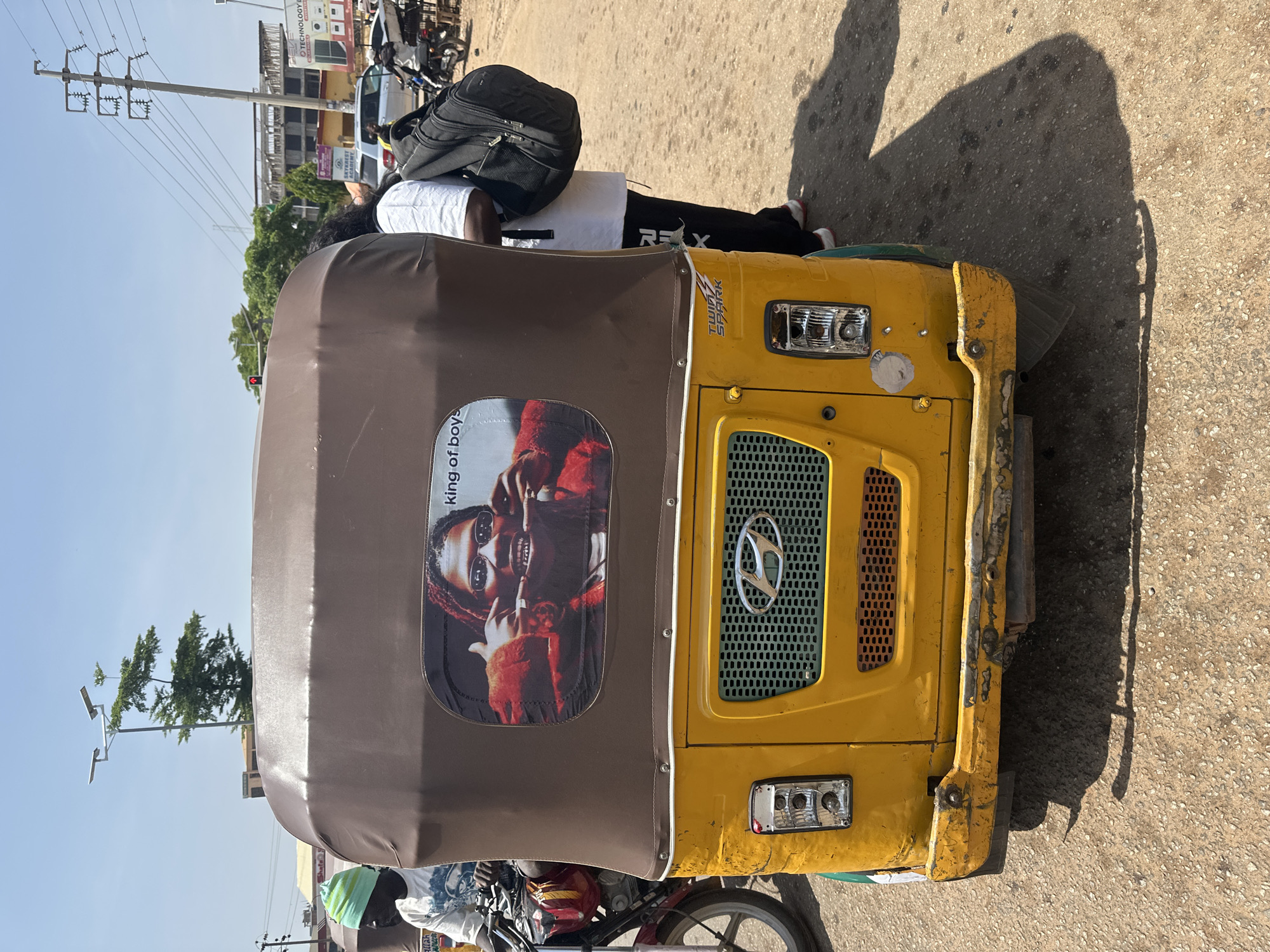 The image shows the rear of a yellow auto-rickshaw parked on a dirt road in Abuja, Nigeria. A person wearing a green beanie and a white t-shirt is visible on the left, seated on a motorcycle parked next to the auto-rickshaw. The auto-rickshaw has a brown canopy with a picture of a person with dreadlocks and sunglasses smiling, and the text "king of boys" printed on it. A person in a white t-shirt and black pants is standing behind the auto-rickshaw, carrying a black backpack. In the background, there are buildings, power lines, streetlights, trees, and other vehicles, including motorcycles and cars, suggesting a busy urban environment. The overall scene depicts a typical street view in Abuja.