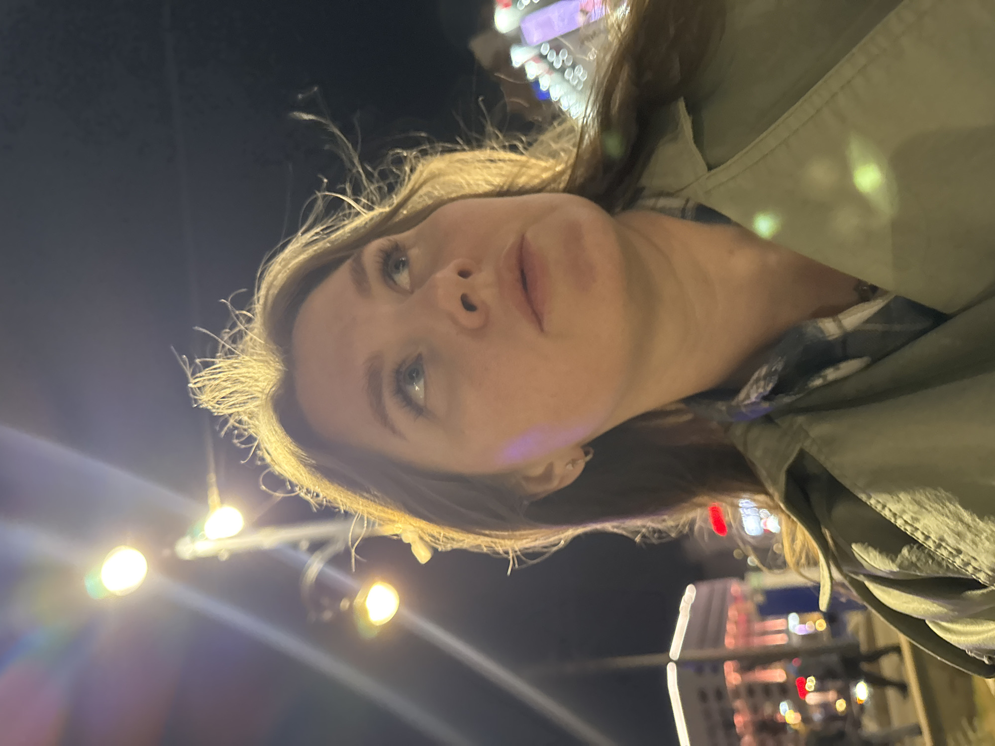 A woman with light skin and light brown hair is taking a selfie outdoors at night. She is wearing a light green jacket. In the background, out of focus, are lights and structures, including what appears to be a Ferris wheel, possibly indicating an amusement park or festive area. Given the location, it could be a popular gathering spot in Perm, Russia, though this cannot be definitively confirmed from the image alone.