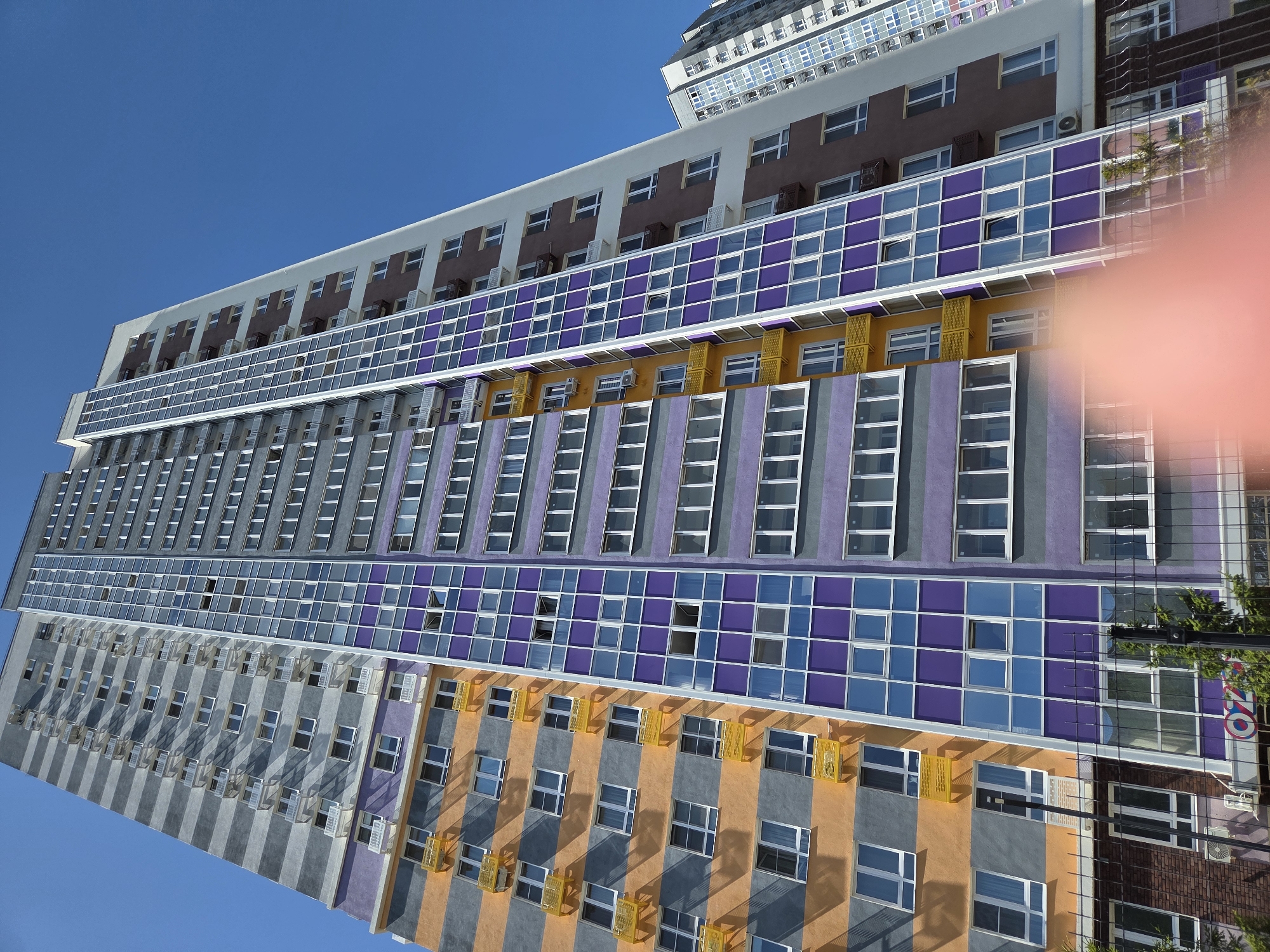 An extreme low-angle shot captures a modern, multi-story apartment building in Ulyanovsk, Russia. The building features a vibrant color scheme with horizontal bands of orange, grey, and purple. The facade is punctuated by numerous windows and balconies, many with white railings. A prominent section of the building is clad in purple and blue glass panels. To the right, another taller apartment building with a more subdued grey and white facade is partially visible. The sky above is a clear, bright blue. The foreground shows a black metal fence and some greenery, with a blurred, pink object, possibly a hand, obscuring a portion of the lower right corner of the image.
