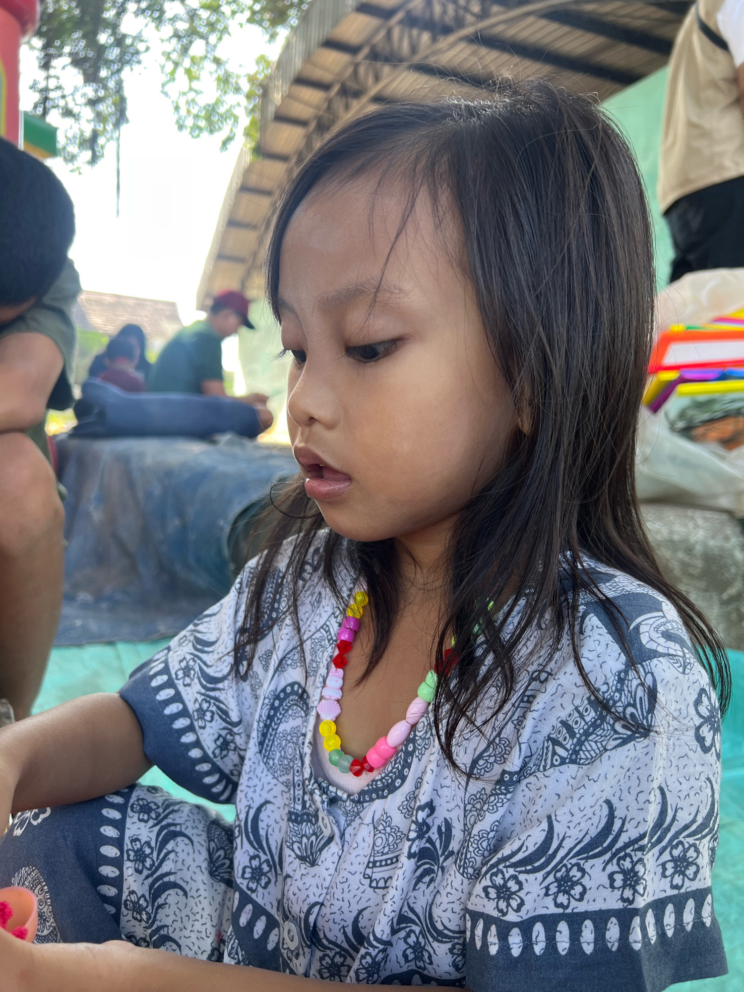In Kecamatan Pati, Indonesia, a close-up, low-angle shot focuses on a young girl of East Asian descent. She is looking downwards and to her right, with her mouth slightly open. Her dark, long hair is partially obscuring her face. She is wearing a white garment with a blue floral and paisley pattern, and a colorful beaded necklace. Her left arm is bent, and her hands are out of frame, though a small, pink object is visible near her hand. In the background, out of focus, are other people sitting or lying down. One person in the mid-ground wears a dark red cap and green shirt, and another person to the far left wears a black beanie. The scene appears to be outdoors, possibly under a makeshift shelter with a corrugated roof, and a blue tarp is visible.