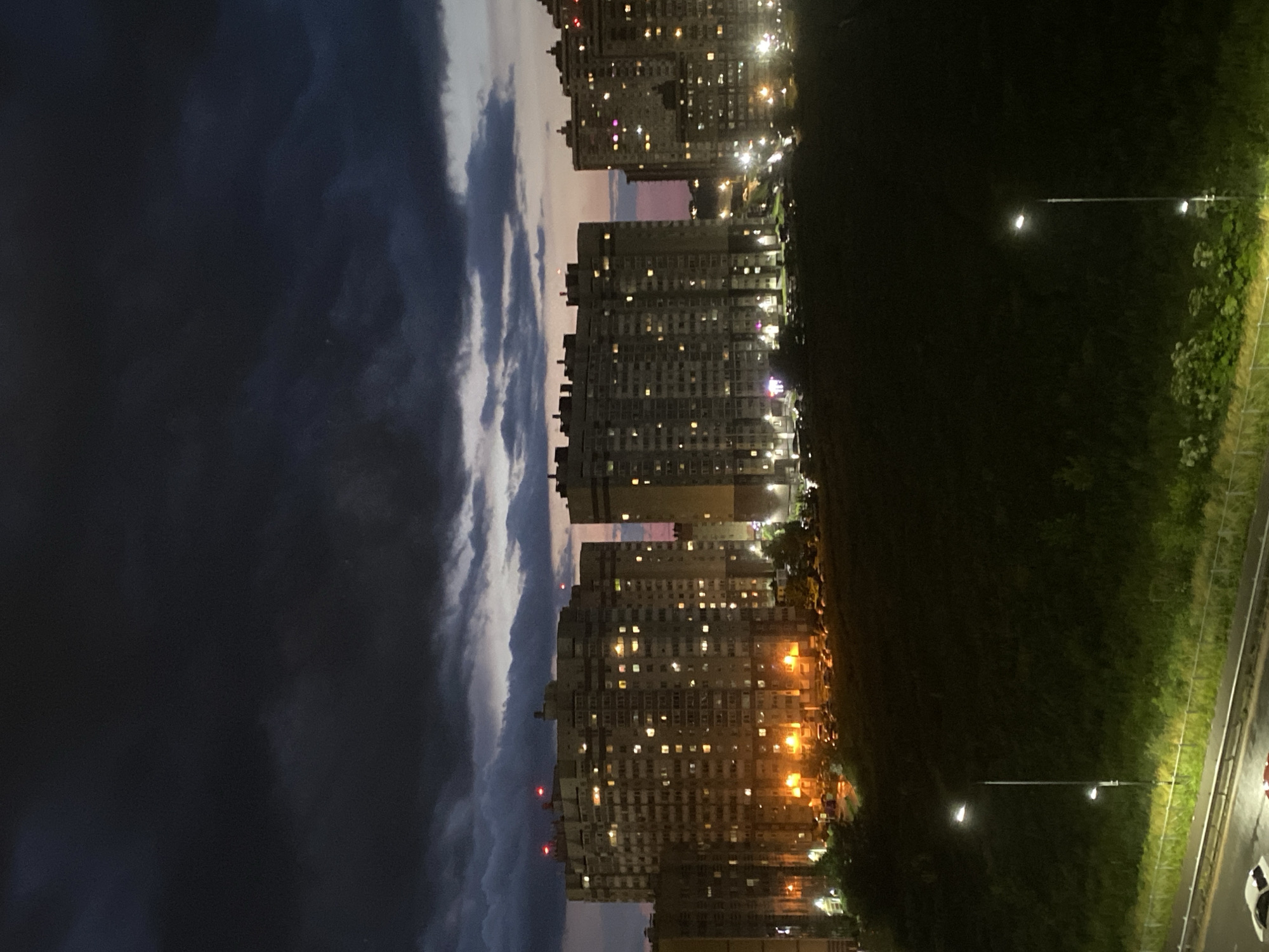 The image captures a twilight scene in the city of Ladozhskoe Ozero, Russia, showcasing a dense urban landscape against a dramatic sky. Dominating the mid-ground are several multi-story apartment buildings, their windows illuminated with warm yellow light, suggesting occupancy and evening activity. The buildings appear to be constructed of concrete and glass, typical of Soviet-era architecture. Red aviation lights are visible atop some of the taller structures.
In front of the buildings, a dark, overgrown hill or embankment slopes downwards, partially obscuring a parking lot filled with numerous cars. The foreground features a dimly lit road or highway with at least two vehicles, one white and one red, partially visible. Streetlights with glowing white lamps cast an eerie illumination on the vegetation and the road. The sky above is a turbulent mix of dark, heavy clouds with hints of a pinkish-orange sunset hue peeking through at the horizon. The overall atmosphere is one of a quiet, encroaching night in a residential urban area. There are no people clearly visible in the image. The scene is static and devoid of any explicit action.