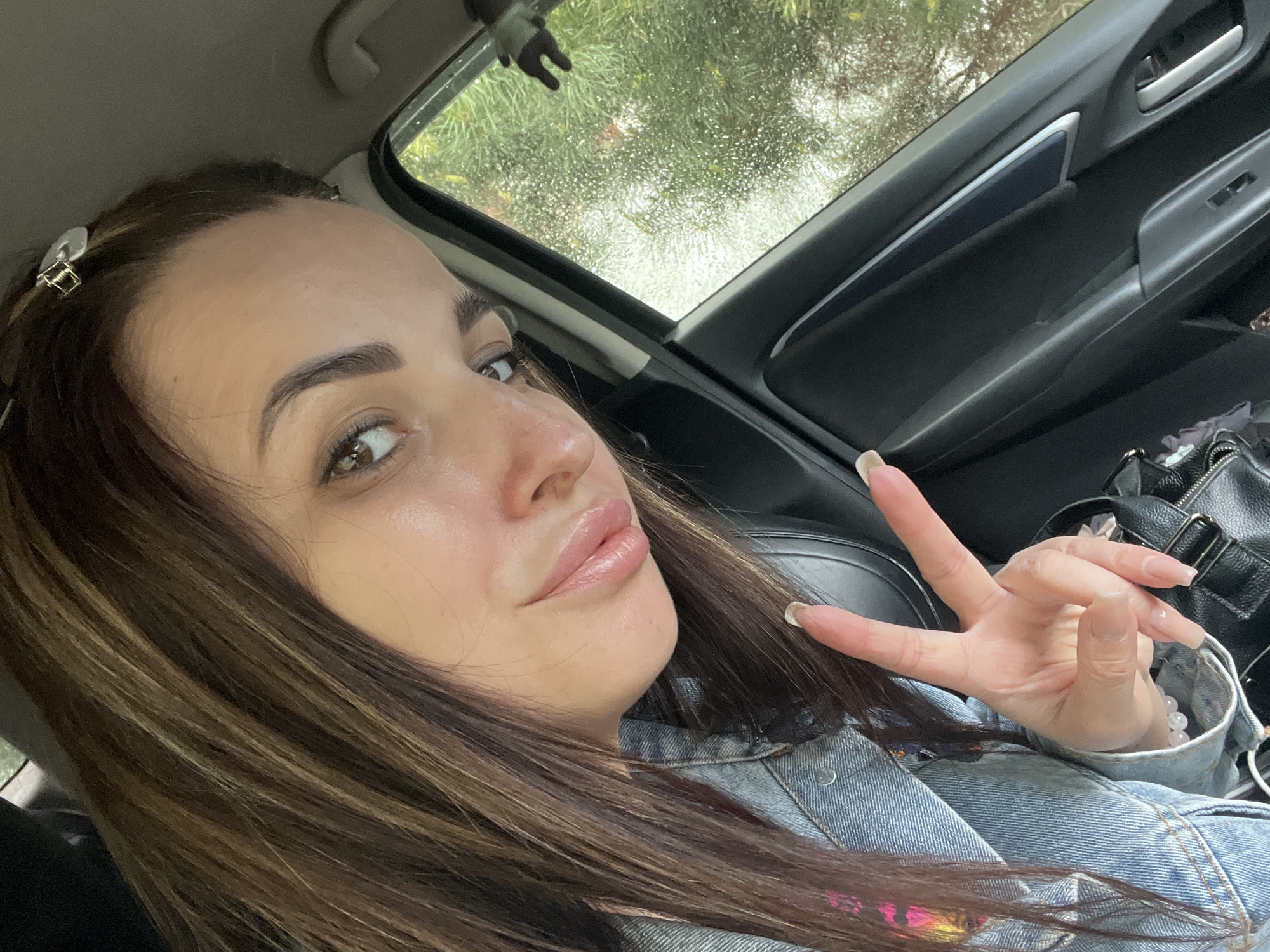 A woman is sitting in a car on a rainy day in the city of Unknown, Unknown. She is wearing a blue denim jacket and has long brown hair with blonde highlights. She is making a peace sign with her right hand. A black purse is visible behind her hand. A silver hair clip is in her hair. Raindrops are visible on the car window.
