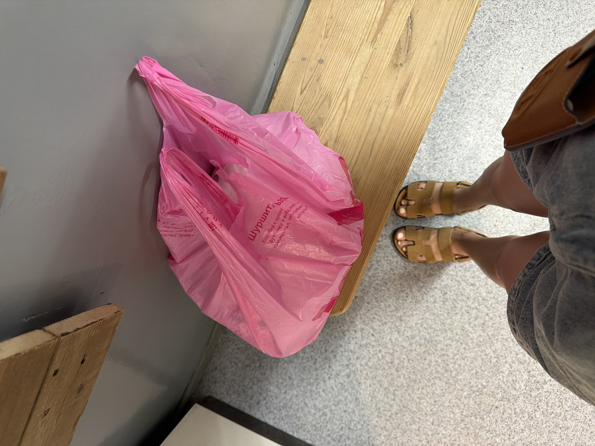 The image shows a first-person perspective of a person standing on a speckled gray floor next to a light-colored wooden bench. The person is wearing denim shorts and tan-colored sandals. To the left of the person is a bright pink plastic bag with some Russian text printed on it. The bag appears to be full and is resting on the bench. In the background, a gray wall is visible, with a wooden shelf mounted high on the wall. The overall scene suggests a casual indoor setting, possibly a changing room, storage area, or a public facility. The location is identified as Semiluki, Russia.