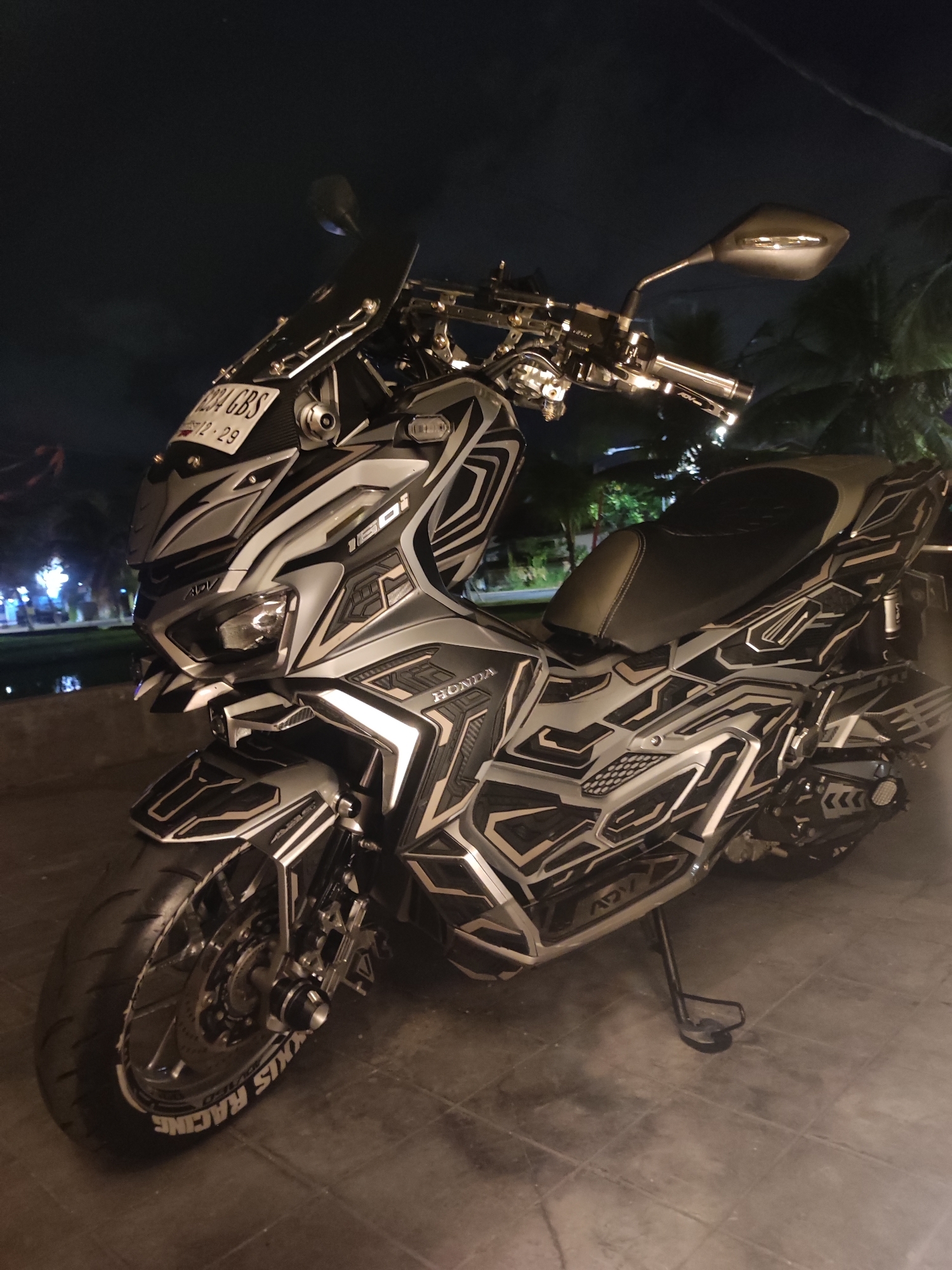 The image depicts a customized Honda ADV 160 motorcycle parked on a tiled surface, likely in North Kuta, Indonesia, given the plate number. The motorcycle features a silver and black geometric design, ADV lettering, and branding on the front tire. The surrounding environment is dimly lit, suggesting it is nighttime. Trees are visible in the background and the edge of water can be seen in the distance. The motorcycle is parked on a kickstand.