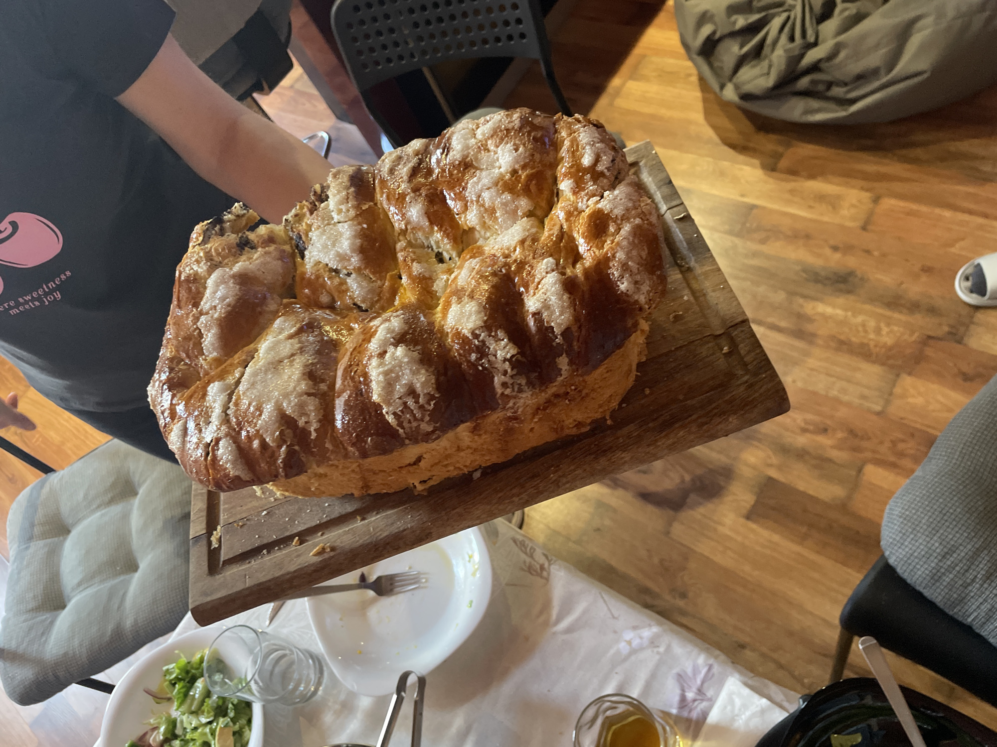 A person holds a large braided sweet bread on a wooden cutting board over a dining table. A black t-shirt with a pink cherry graphic is visible on the person holding the bread. The table is covered with a white tablecloth. There are plates, a fork, a glass of orange liquid, and a bowl of salad on the table. The background includes wooden floors, chairs, and a beanbag. The location in Burgas, Bulgaria is not evident in the image.