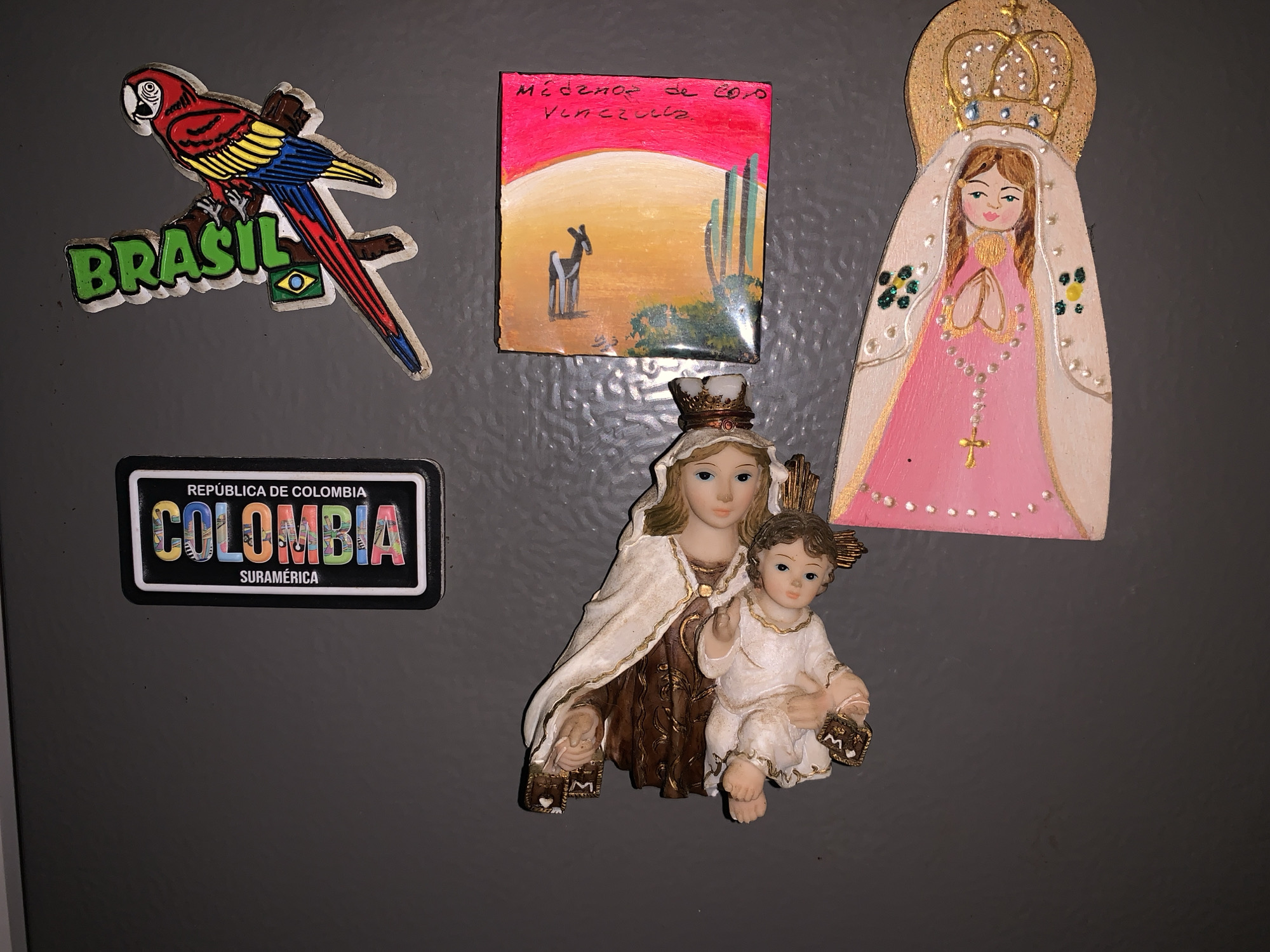 The image depicts a collection of magnets affixed to a gray surface. The magnets include a parrot with "Brasil" text and a Brazilian flag, a rectangular magnet with the words "República de Colombia" and "Colombia Suramérica," a depiction of "Médanos de Coro, Venezuela," and two religious-themed figures, one representing the Virgin Mary with a rosary, and the other showing Mary with Jesus. No location of Santa Ana is visible in this image.