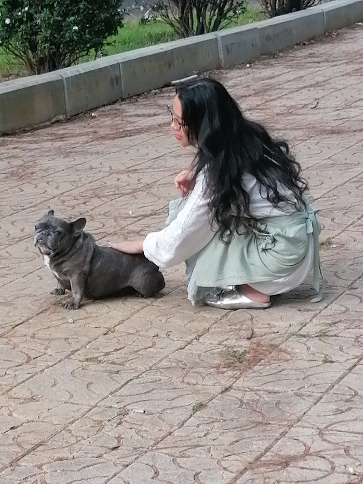 The image depicts a woman and a French bulldog in an outdoor setting, likely a park or public walkway in Da Lat, Vietnam. The woman, with long dark hair and glasses, is squatting down, wearing a white top and a light green skirt or apron. She has her right hand gently resting on the back of the dog. The French bulldog is a dark gray color with a white marking on its chest, and it is sitting down, looking slightly towards the camera. The ground is paved with light-colored tiles, some of which have a circular pattern. In the background, there is a low concrete wall, followed by greenery and trees, consistent with the natural landscape found in Da Lat. The overall lighting suggests it is daytime.
