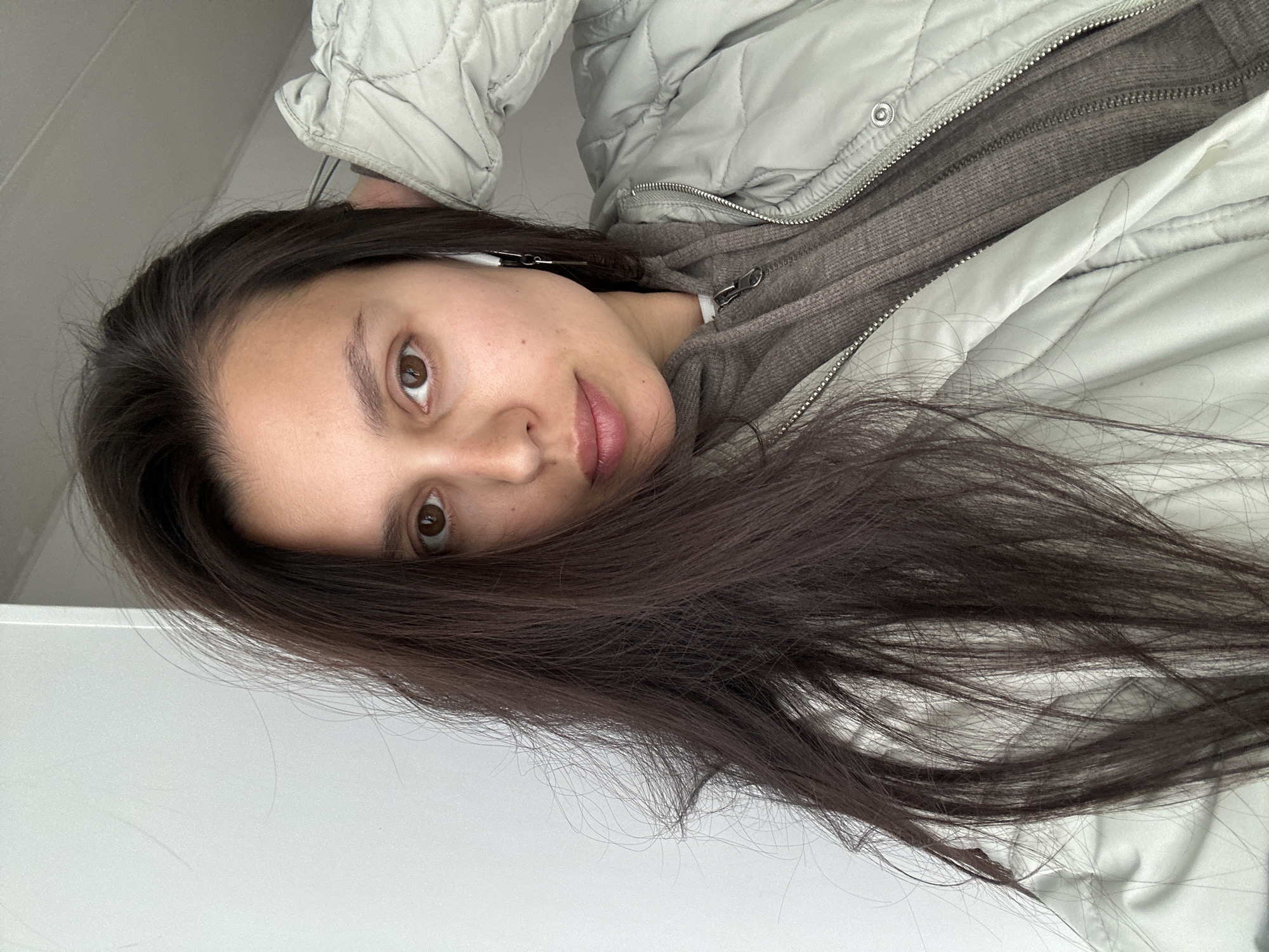 A young woman with long, dark brown hair is wearing a gray quilted jacket and a brown zippered hoodie. She is also wearing an earbud. The background is white, likely a wall. There is no evidence of the city of Chaikovsky, Russia.