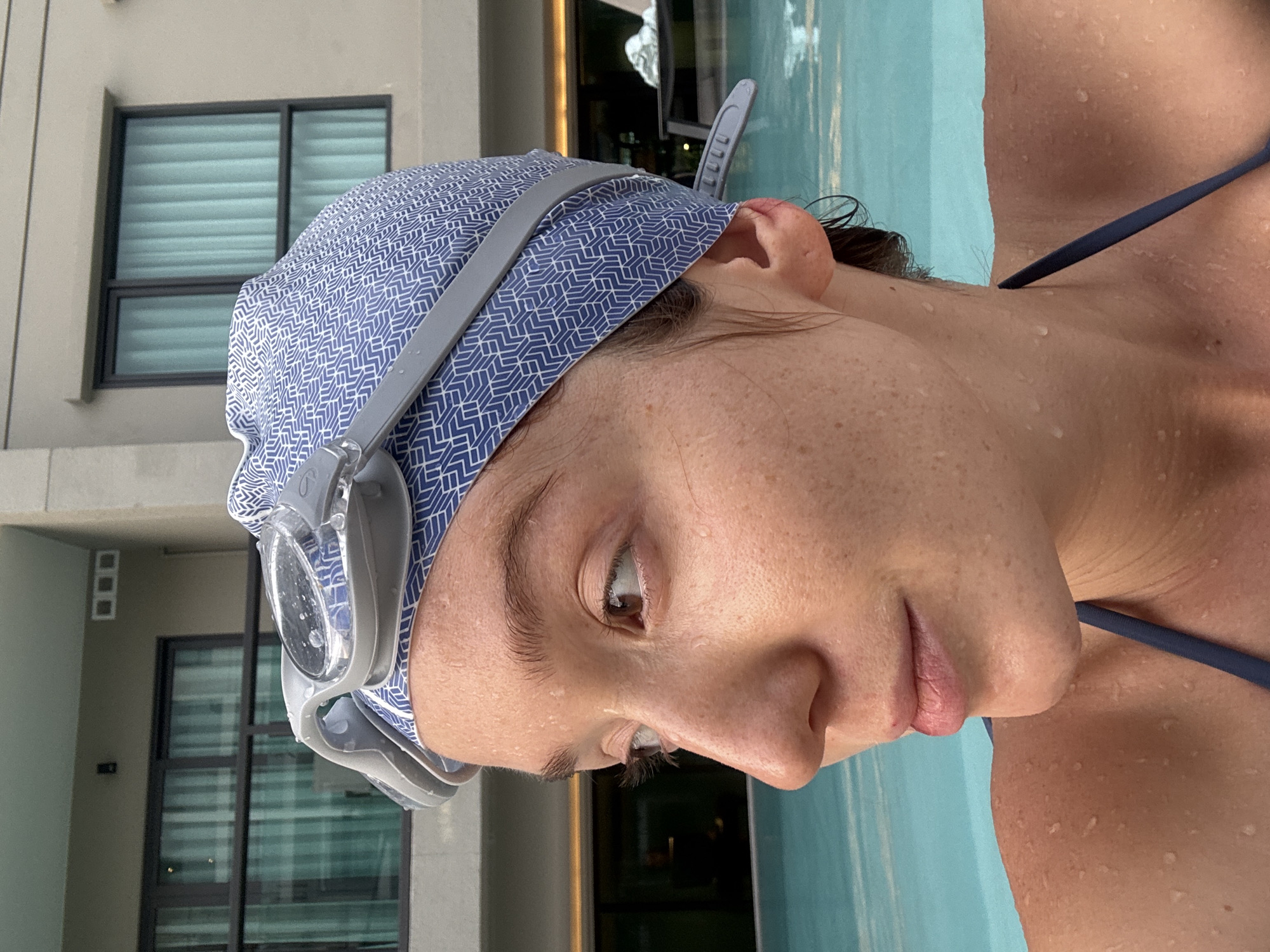 The image shows a close-up, angled portrait of a person in a swimming pool setting. The individual, appearing to be a woman, is wearing a blue and white patterned swim cap and grey swimming goggles, which are resting on the cap. Water droplets are visible on her face, neck, and shoulder, indicating she has recently been swimming or is wet from the pool. She has dark hair visible around her ears and under the cap. Her expression appears calm and contemplative as she looks slightly downwards and to her right. A dark blue swimsuit strap is visible on her shoulder.
In the background, a light-colored building with large, dark-framed windows is visible. The windows feature horizontal blinds. The vibrant blue water of the pool occupies the right side of the frame. The scene conveys a relaxed moment by a swimming pool, likely during the daytime. Given the context, this setting is in Choeng Thale, Thailand. No visible text is present in the image.