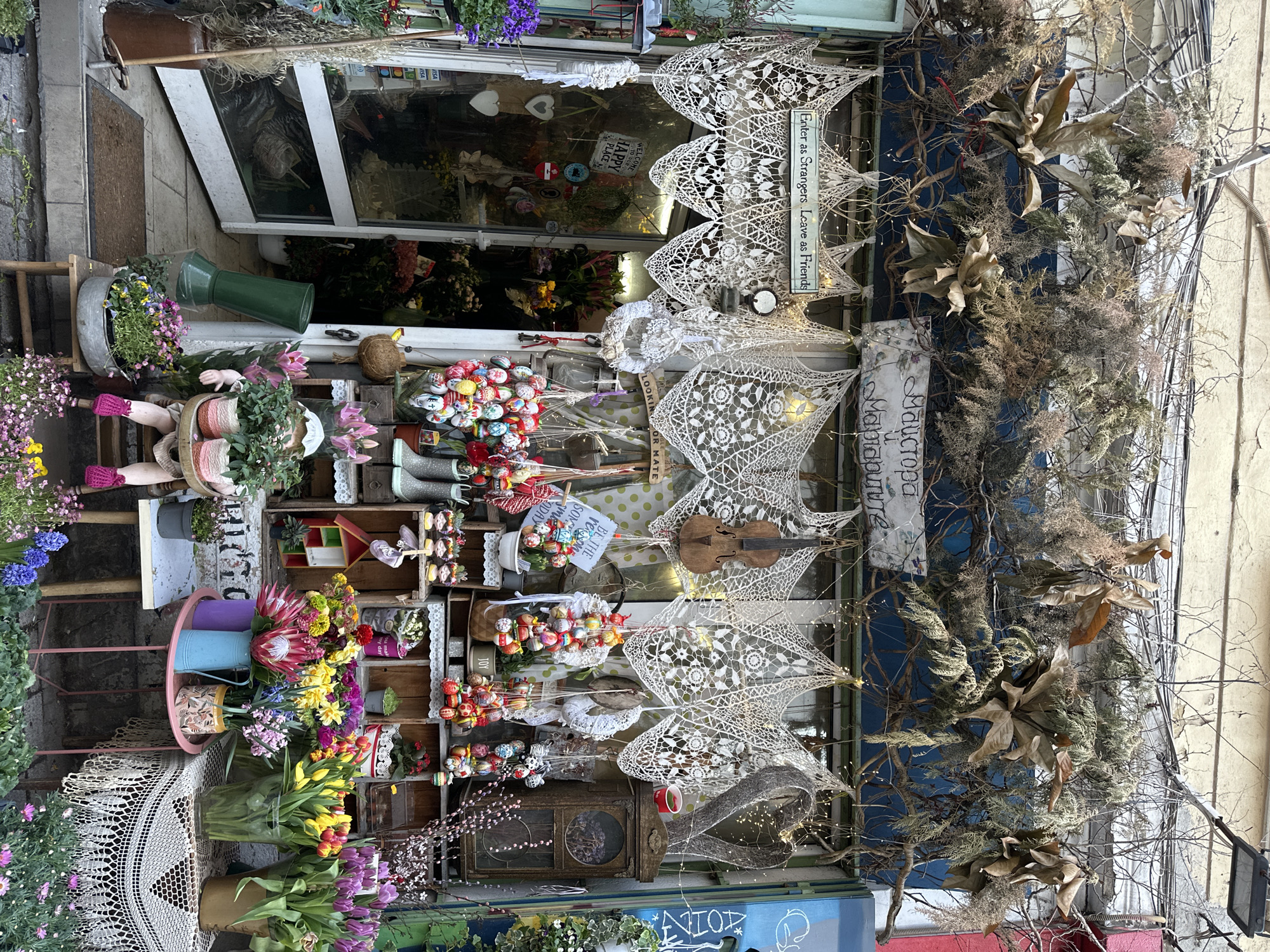 The image shows a storefront in Sofia, Bulgaria. The shop is adorned with an eclectic mix of decorations: white lace doilies hang in rows, interspersed with dried branches, vintage radios, violins, and strings of lights. Various arrangements of flowers, including tulips, hyacinths, and proteas, are displayed in buckets and wooden crates. A sign reads "Enter as Strangers, Leave as Friends" and "Welcome to our happy place". There are signs in cyrillic as well, one reads "Майстора Мабиапури", which likely is the name of the shop. The overall aesthetic is rustic and whimsical.