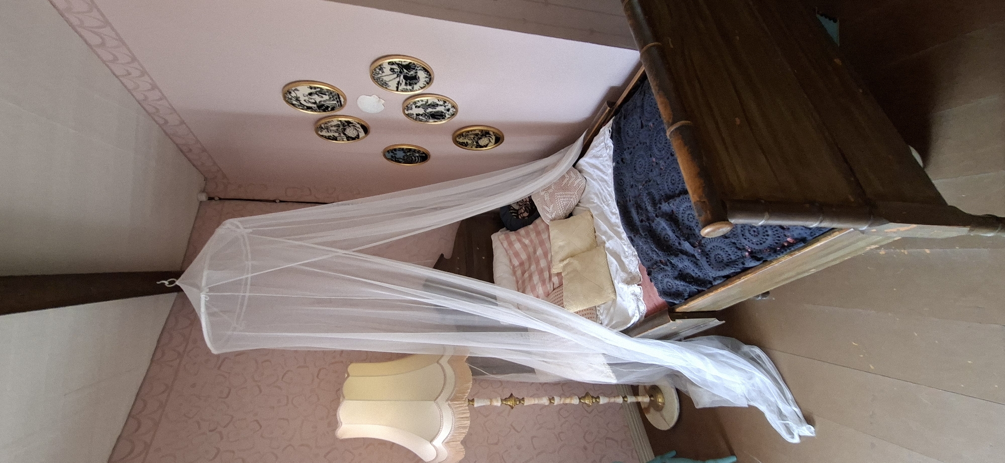 The image shows a bed with a mosquito net draped over it, positioned in a room with pink walls. A floor lamp with a tasseled shade stands to the left of the bed. On the wall above the bed, a collection of six decorative plates with black and white scenes is displayed. The bed is covered with a dark blue blanket and several pillows. The flooring appears to be wooden planks. There are no people visible in the image. The scene appears to be an interior bedroom setting. The location in Naantali, Finland, is not discernible from the visual information provided.