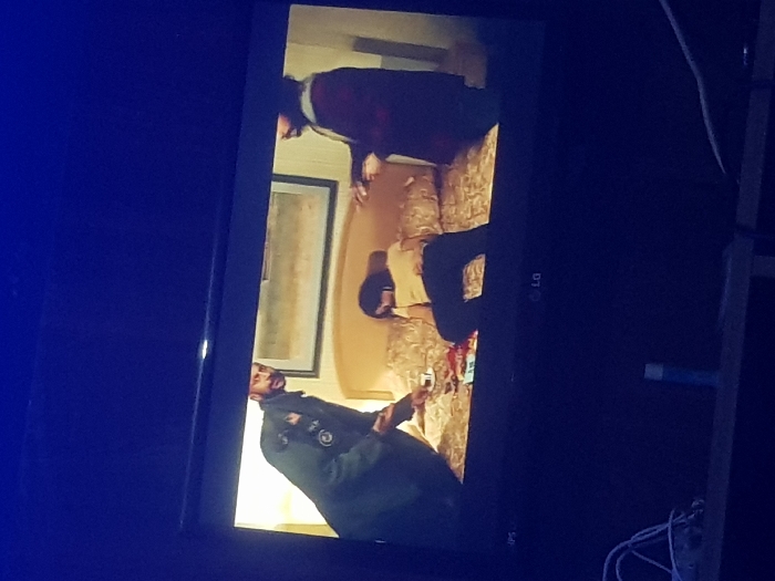 The image depicts a television displaying a scene with three people in a room. A man in a green jacket sits on the edge of a bed, holding a glass. Two other figures, one wearing a red and gray top, are situated on the bed. There is a framed picture on the wall behind the bed. An LG logo is visible on the television frame. Cords are present in the lower right of the shot.