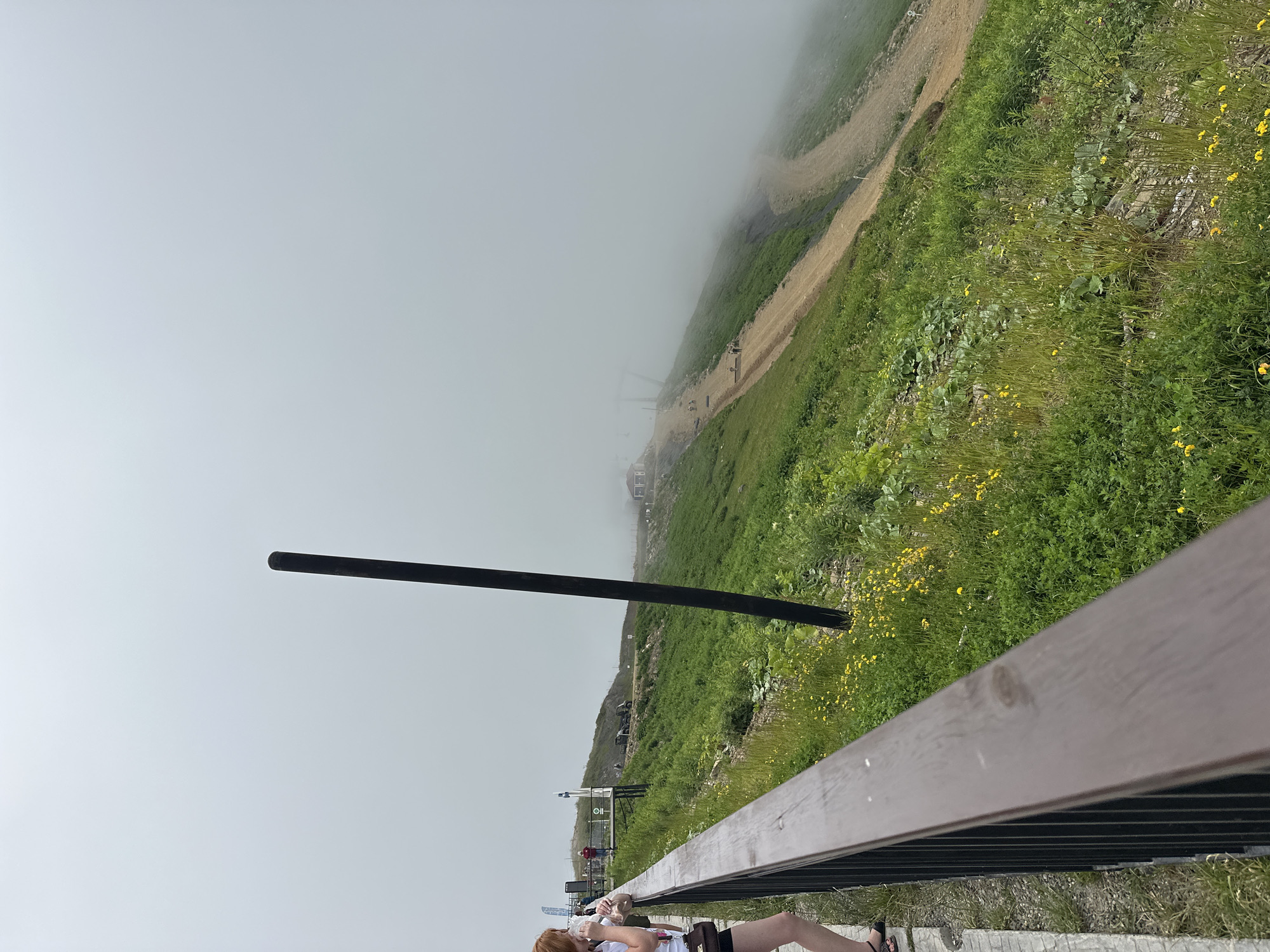 The image captures a view from a wooden railing overlooking a grassy mountainside in Estosadok, Russia. The sky is overcast with thick fog obscuring the upper reaches of the mountain and the distant landscape. A dark, vertical pole stands prominently in the foreground, partially silhouetted against the muted sky. On the left side of the frame, a woman with red hair is visible, wearing a white top and dark shorts, and appears to be holding a drink. She is standing on a paved pathway beside the railing. The mountainside itself is covered in lush green grass and dotted with small yellow wildflowers. A dirt path winds its way up the slope, and several indistinct figures can be seen walking on it. In the distance, a small building with a red roof is visible, partially shrouded by the fog, along with what appears to be a ski lift structure. The overall impression is one of a cool, misty day on a mountain resort.