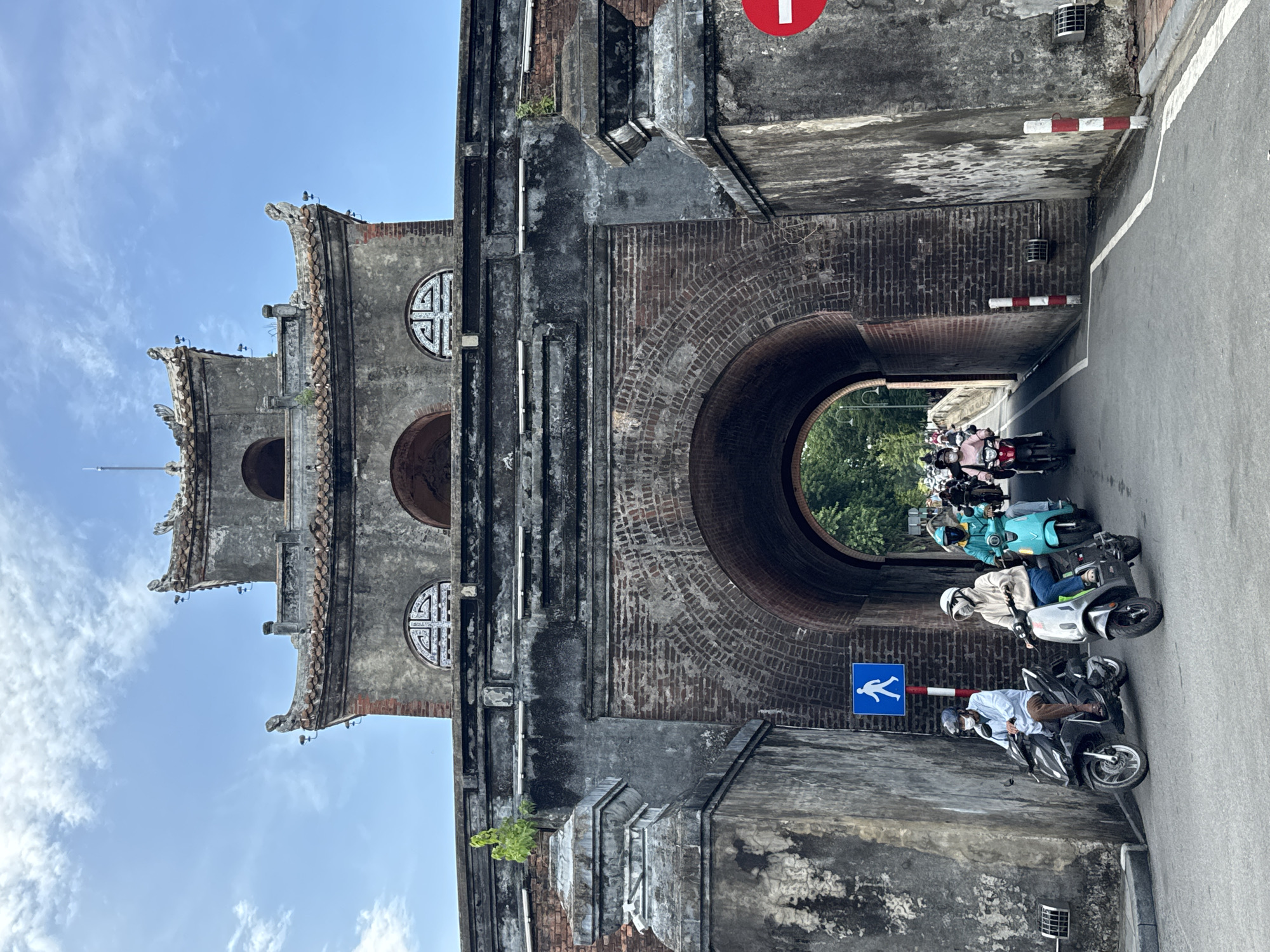 In Phú Xuân Ward, Vietnam, a wide brick archway, part of a historical structure, allows passage for a line of motorbikes. The archway is framed by weathered stone and brick pillars. Above the arch, the building features intricate architectural details, including tiled roofs with decorative elements and ornate window designs. Several individuals on motorbikes are moving through the archway. Two motorbikes are visible in the foreground. One is black, ridden by a person wearing a light blue shirt and brown pants, with a face mask and helmet. The second motorbike is silver, ridden by a person in a light-colored hooded jacket and jeans, also wearing a face mask and head covering. A blue pedestrian crossing sign with a white figure is posted on the left. Beyond the arch, more motorbikes and greenery are visible. The sky above is blue with scattered clouds.