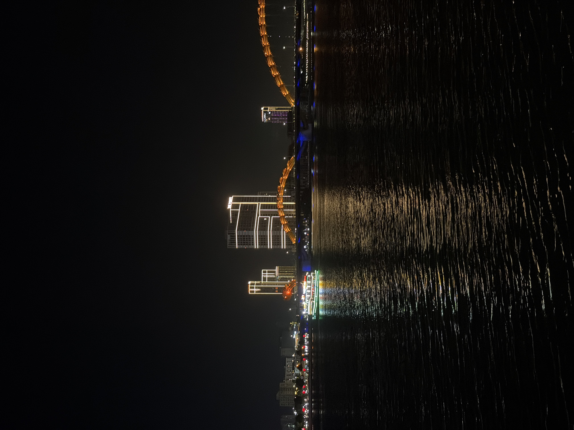The image captures a striking night scene of a city waterfront in Đà Nẵng, Vietnam. The sky is pitch black, providing a dramatic backdrop for the illuminated urban landscape.
In the midground, several modern high-rise buildings line the waterfront, their facades brightly outlined with warm white and yellow lights, creating geometric patterns. A prominent arch bridge, likely the Dragon Bridge, spans across the water, its structure aglow with golden-orange lights. A second, similar bridge is visible further in the distance.
The lower portion of the image is dominated by a dark body of water, probably the Hàn River, which vividly reflects the city lights. These reflections appear as elongated, shimmering streaks of light, indicating a gentle rippling of the water's surface. On the water, closer to the foreground, an illuminated tourist boat is visible, adorned with bright green, red, and white lights. Beyond the main structures, countless smaller lights extend along the distant shoreline, suggesting a sprawling cityscape.
The scene conveys the vibrant and picturesque atmosphere of Đà Nẵng at night, characterized by architectural illumination and reflections on the water. There is no visible text in the image.