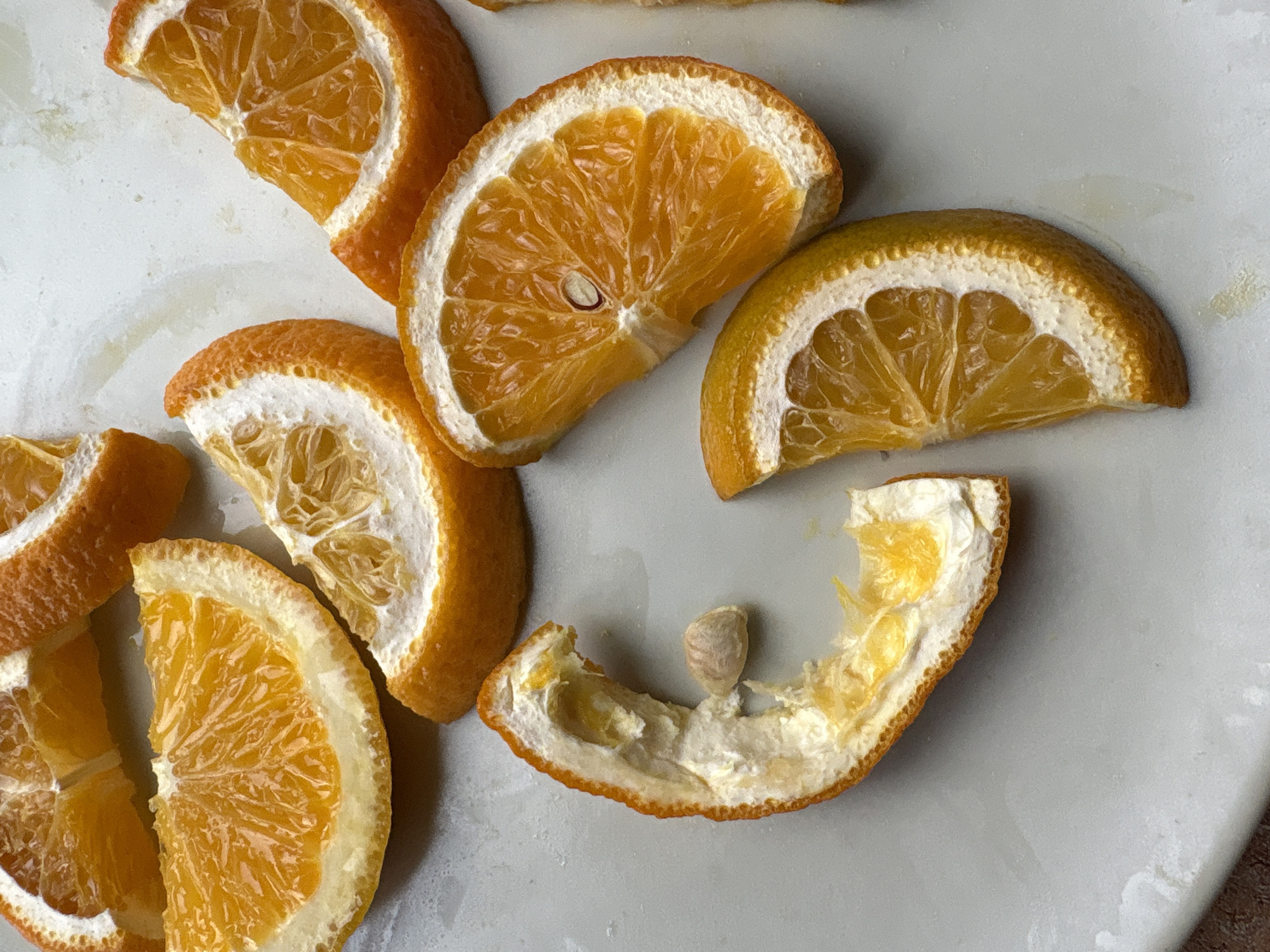 A close-up, overhead shot captures several slices of orange arranged on a pale gray, textured surface, possibly a plate or countertop. The oranges are cut into wedges, revealing their vibrant orange pulp and white pith. Some slices are whole, while others appear to be partially dried or shriveled, with the pith more prominent. One slice stands out as a hollowed-out piece of rind, with a single seed resting inside. The lighting is bright, creating highlights and shadows that emphasize the texture of the fruit. The background is out of focus, suggesting a shallow depth of field. The scene is devoid of any human presence or activity. No specific location in Vitsebsk, Belarus can be determined from the provided image.