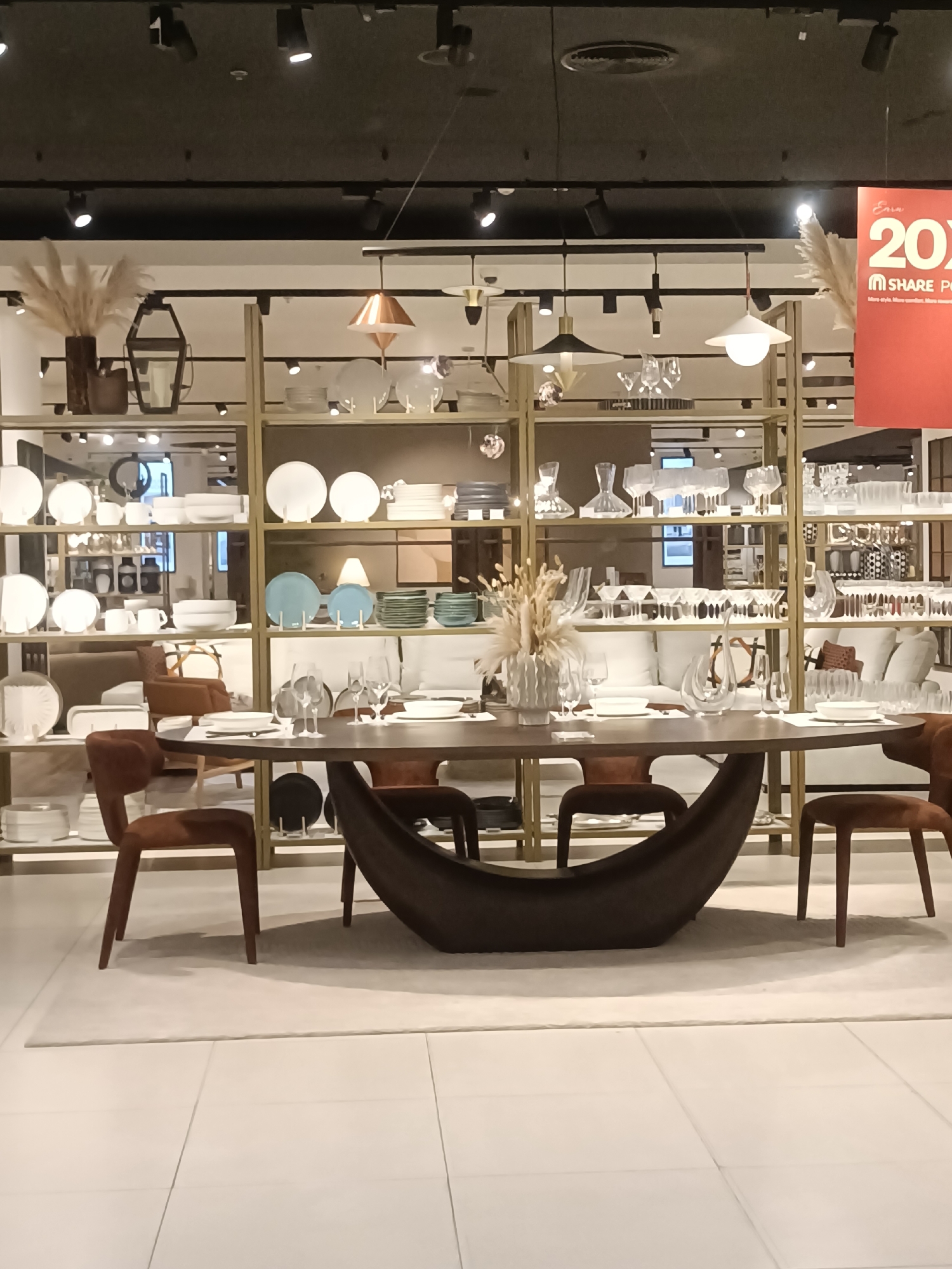 The image shows a retail display of home goods, likely in a store located in Dubai, United Arab Emirates. The scene features a large oval dining table with several chairs around it, set with plates and glassware. Behind the table, a tall shelving unit displays an assortment of dinnerware, decorative vases, and lanterns. The background includes more shelving with similar items, as well as what appears to be a living room setup with a sofa and armchairs. A large red promotional banner with the text "20%" is visible on the right side of the image. The lighting is bright, typical of a retail environment.